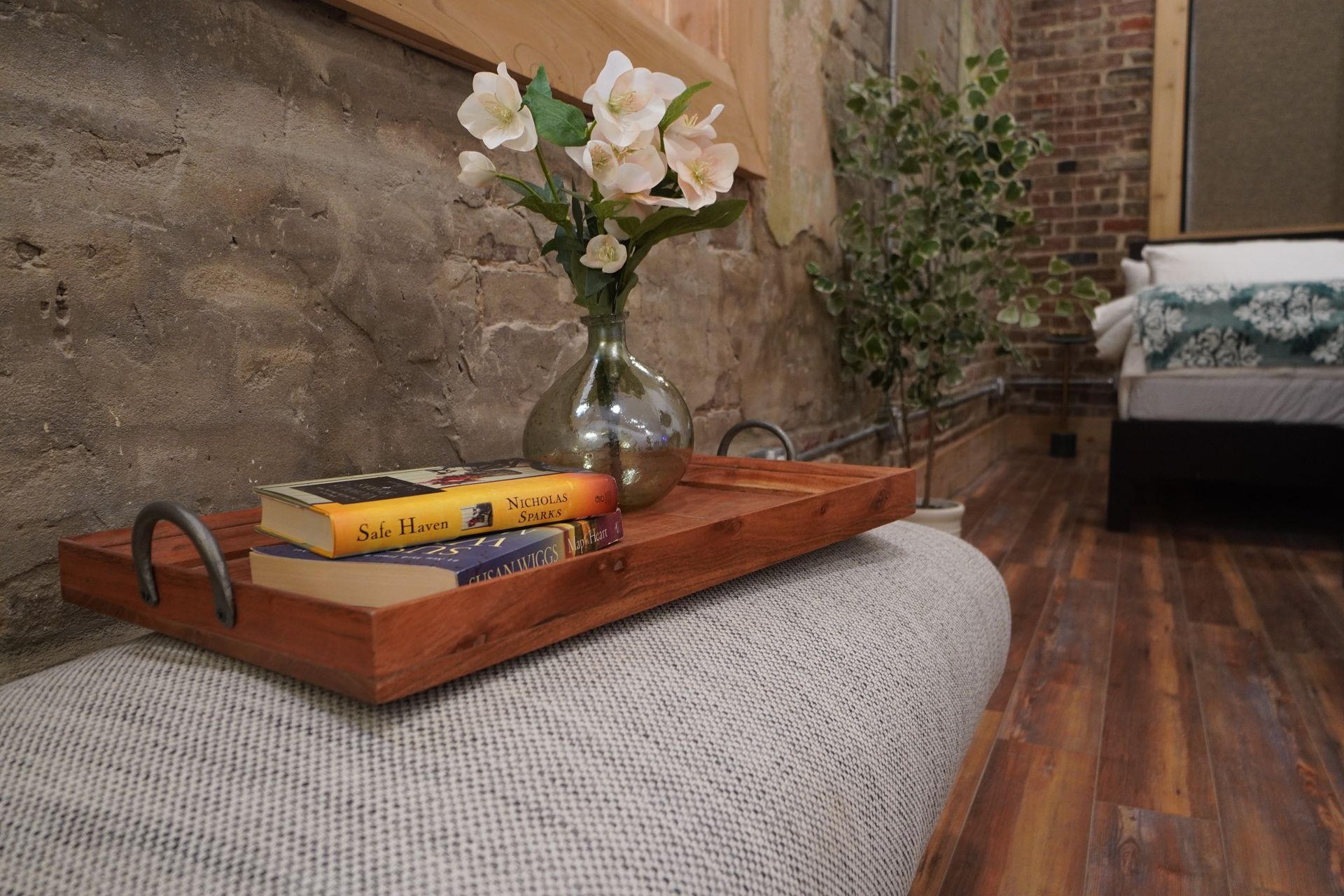 Wooden tray with vase of flowers and books on a gray upholstered bench in a room with a brick wall.
