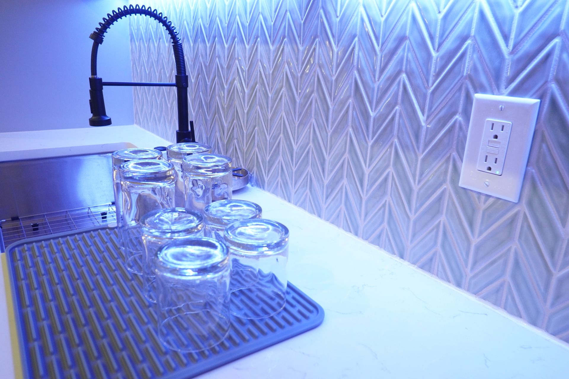 Kitchen countertop with glasses, sink, and backsplash, illuminated with blue light. An electrical outlet is visible.