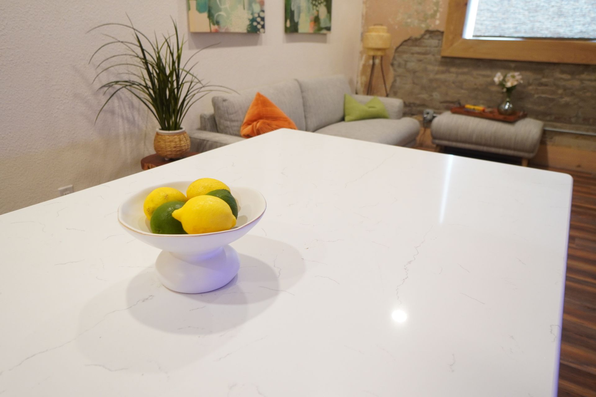 White kitchen island with bowl of lemons, living room with sofa and art in background.