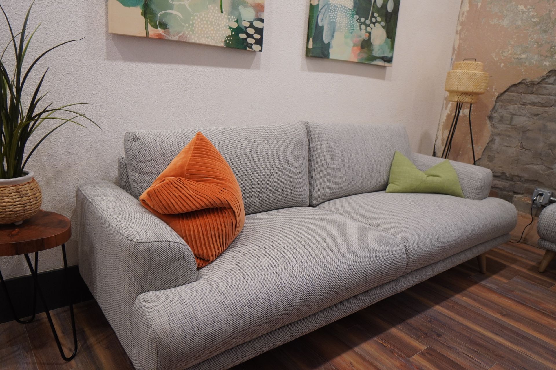 Gray sofa with orange and green pillows, next to a small wooden table with a plant, art on wall.