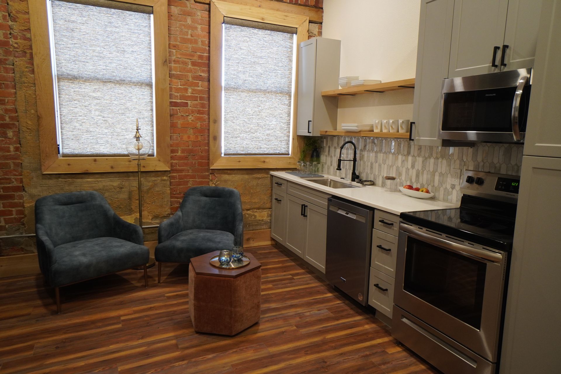Modern kitchen with brick wall, two blue chairs, stainless steel appliances, and wooden floors.