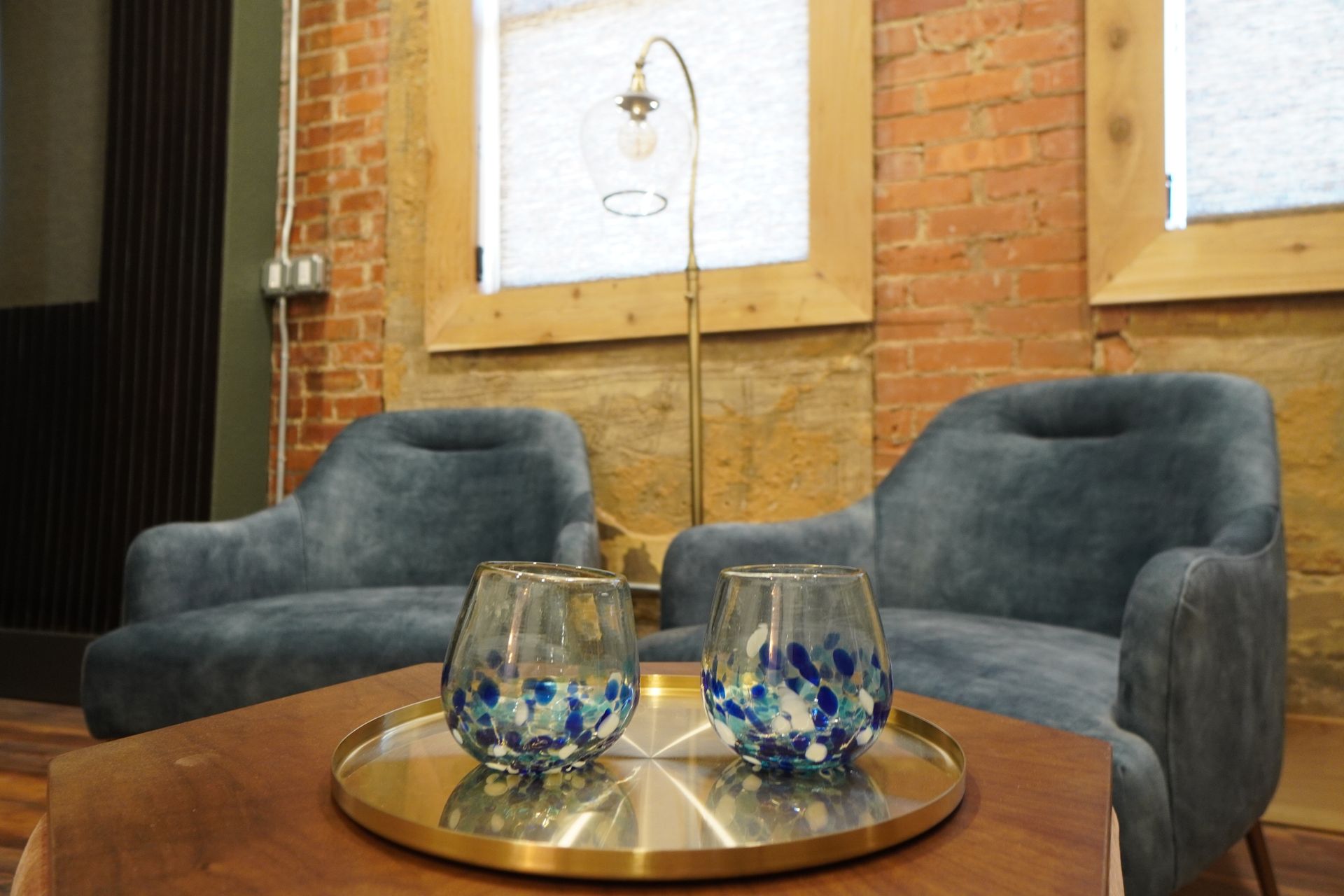 Two blue armchairs in front of a brick wall, with a gold tray holding two decorated glasses on a wooden table.