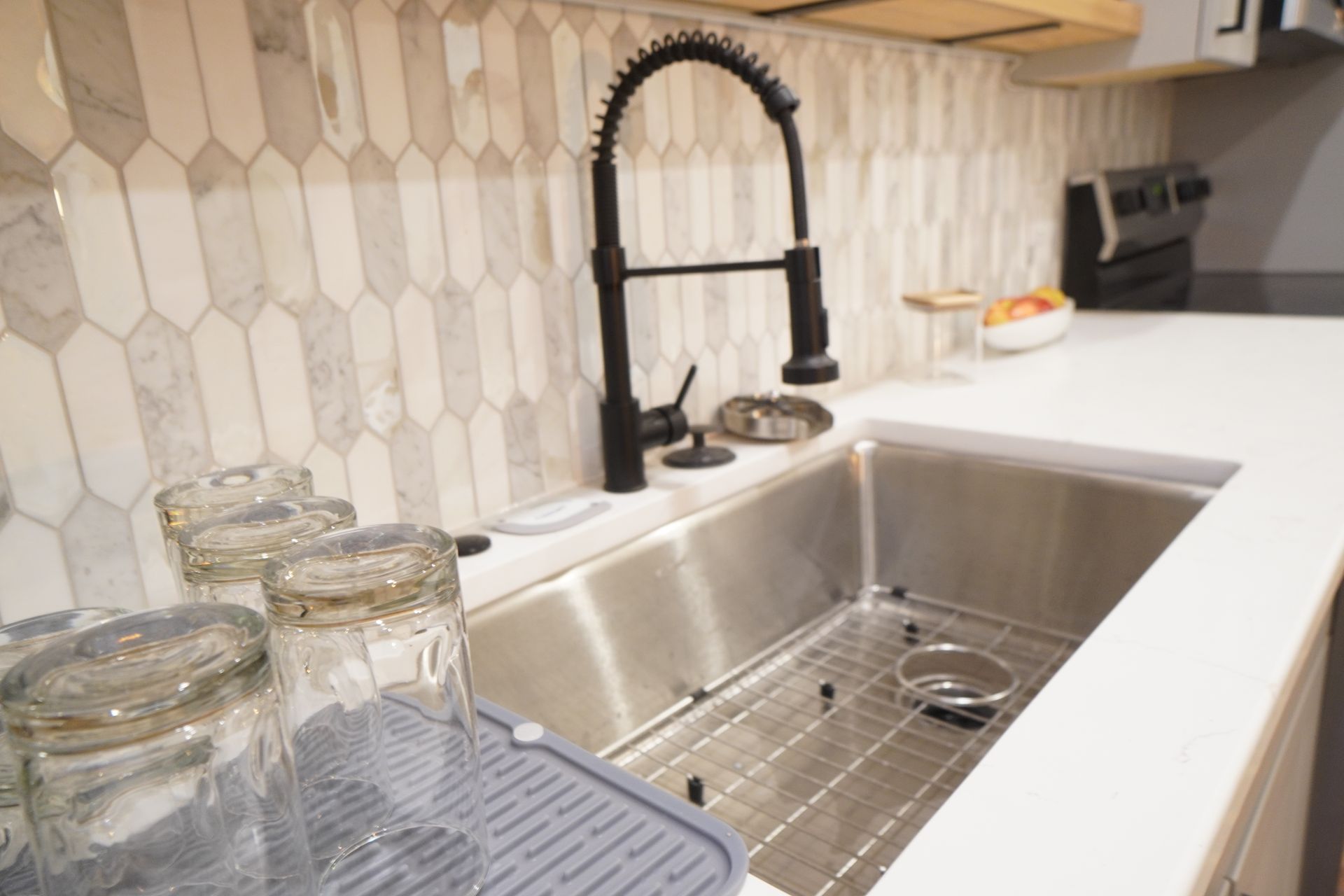 Kitchen sink with black faucet, glasses, and white countertop. Tile backsplash in the background.
