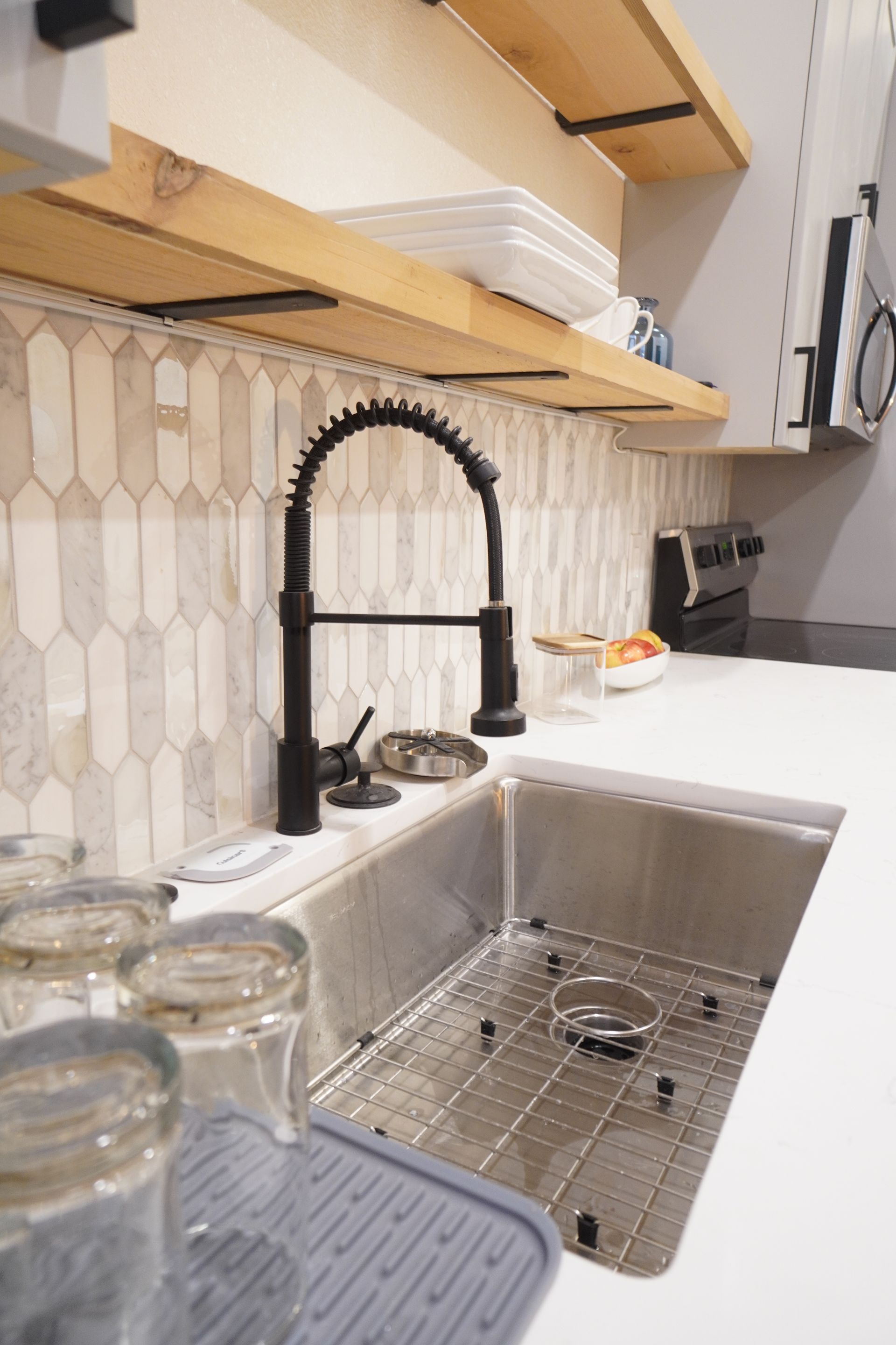 Kitchen sink with black faucet, stainless steel basin, white countertop, and light-colored backsplash tile.