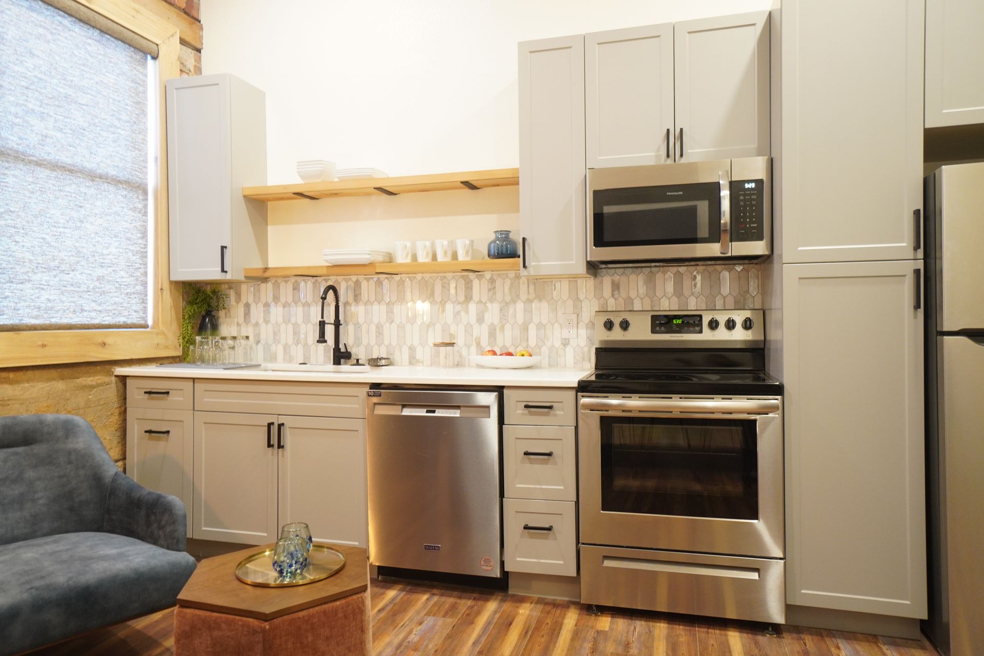 Modern kitchen with stainless steel appliances, light gray cabinets, and a wood accent shelf.