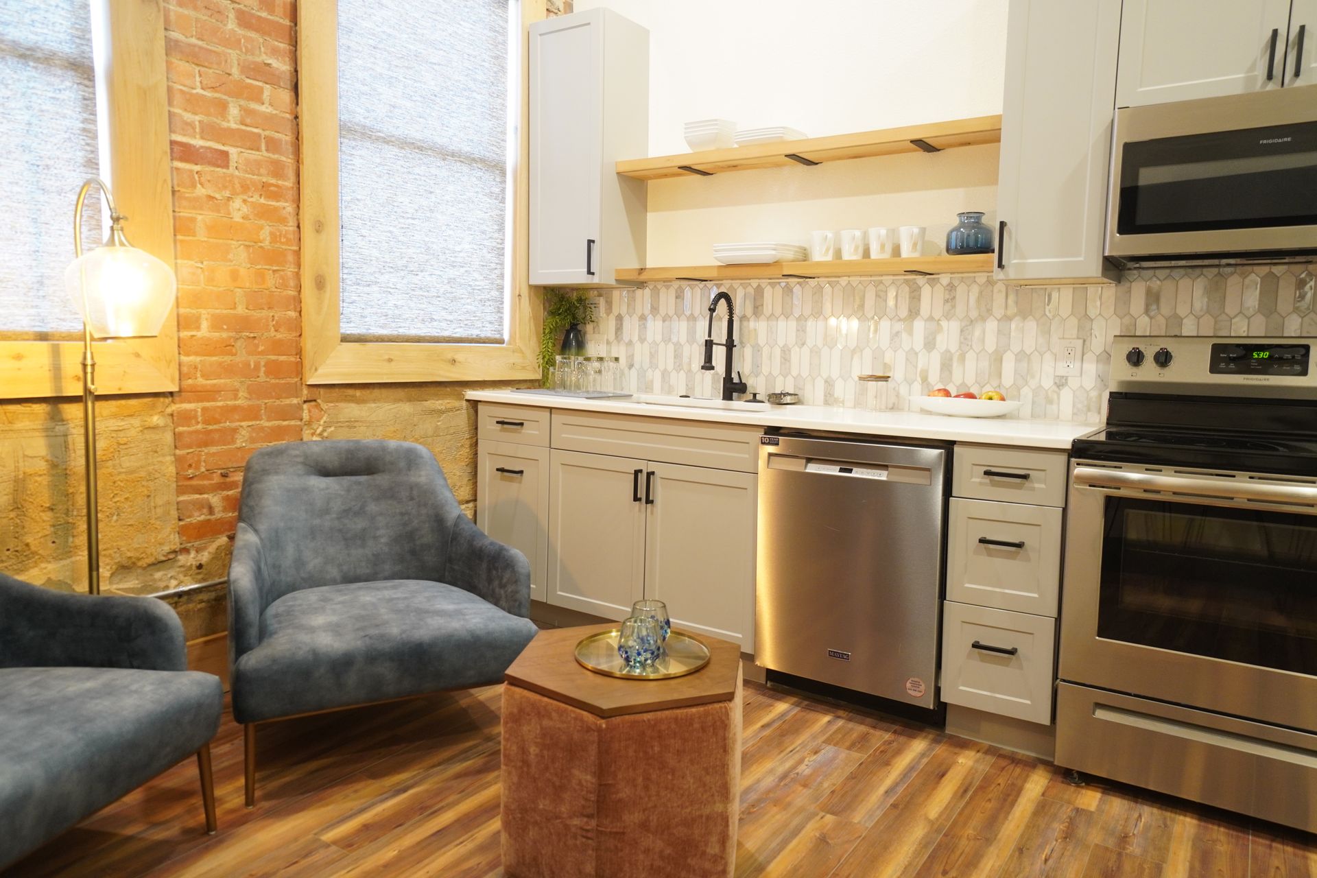 Kitchen with brick wall, light gray cabinets, stainless steel appliances, and two blue armchairs.