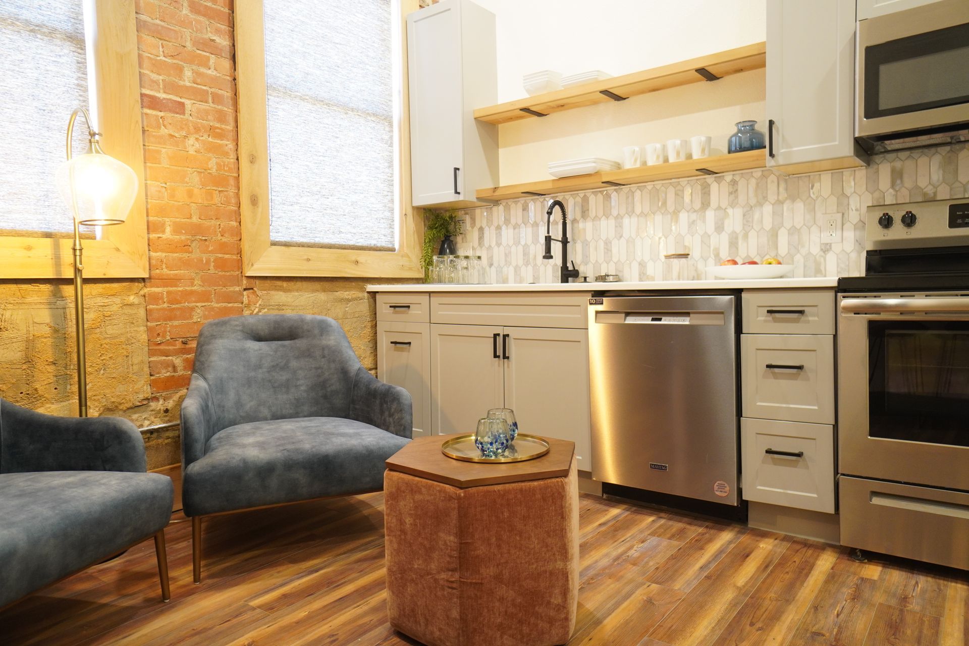 Cozy kitchen with brick wall, light cabinets, stainless steel appliances, and two blue armchairs.