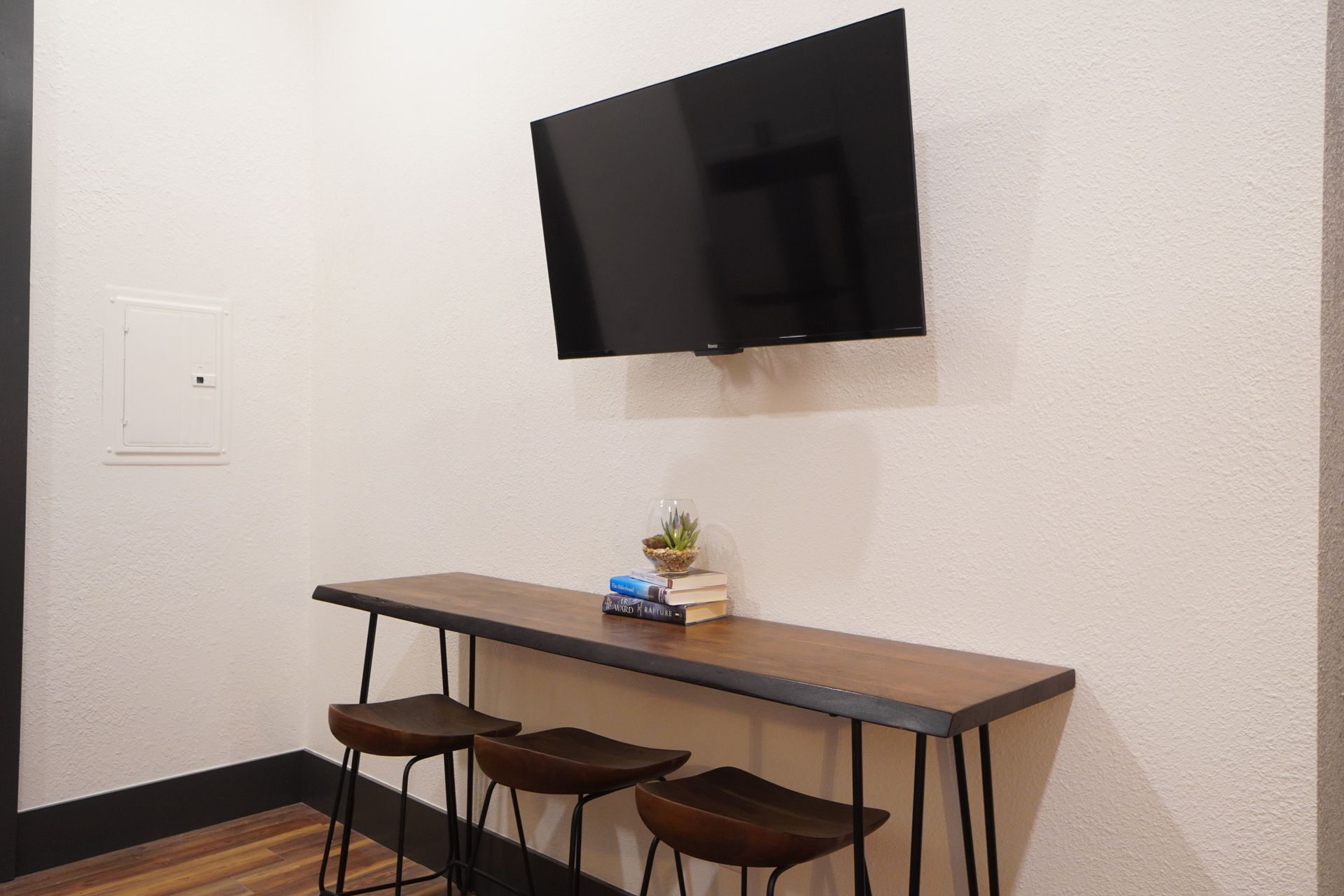 TV above a long, wooden bar with three stools. Succulents and books are on the bar. White walls.