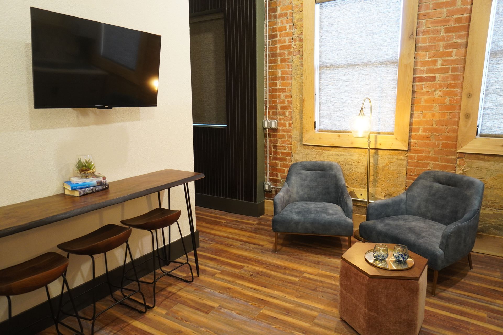 Modern lounge area with a TV, brick wall, two blue armchairs, and a wood bar with stools.
