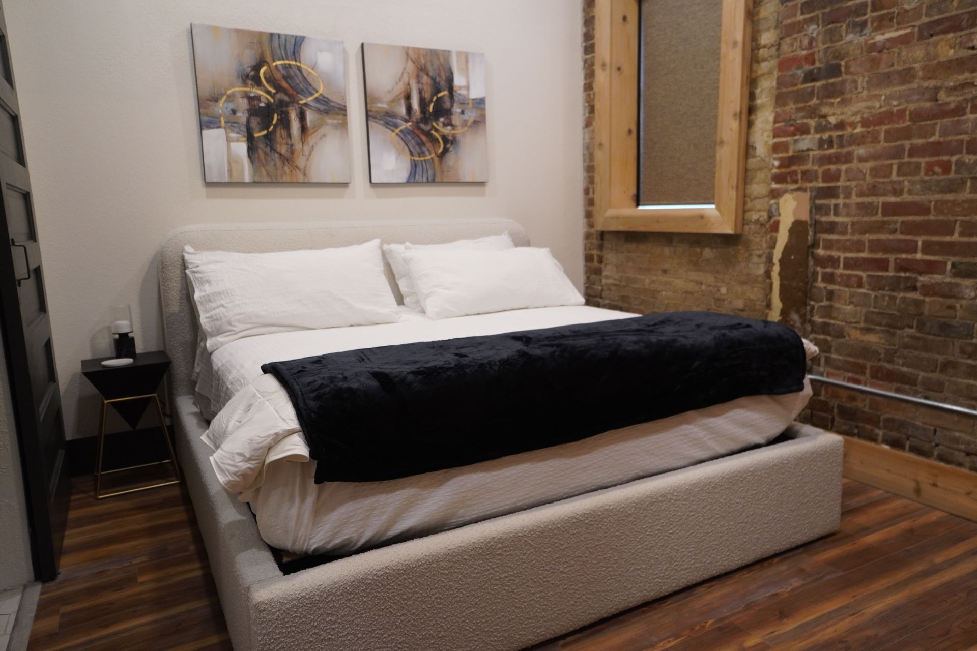 Bedroom with bed, white bedding, black throw, artwork, and exposed brick wall.