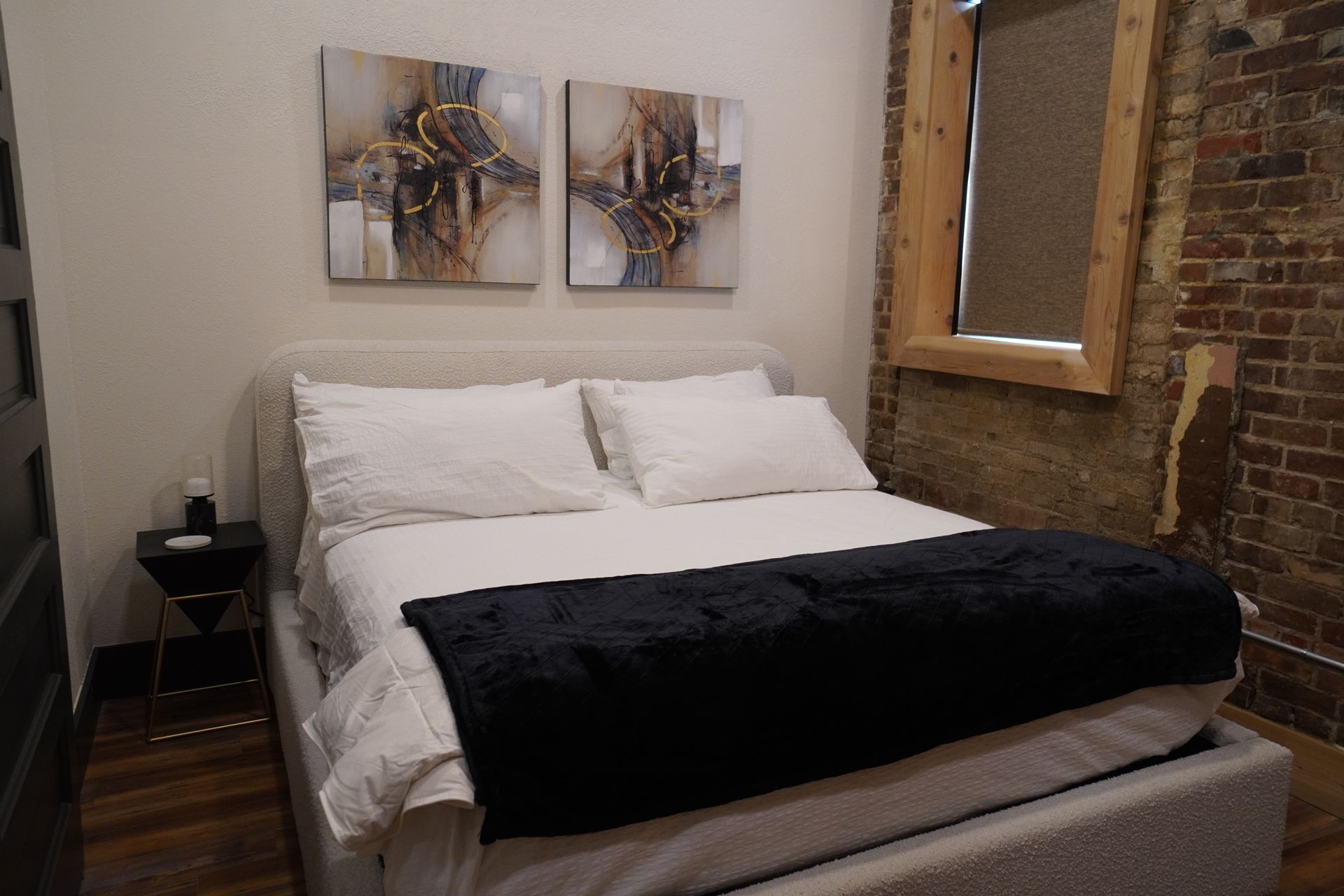 Bedroom with bed, pillows, black throw, artwork, exposed brick, and window.