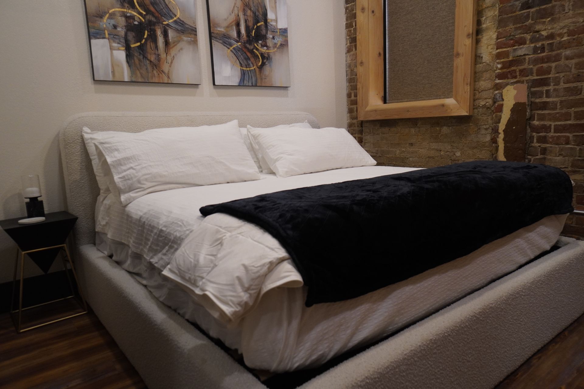 Bedroom with a bed made with white linens and a black throw blanket. There's a bedside table and art on the wall.