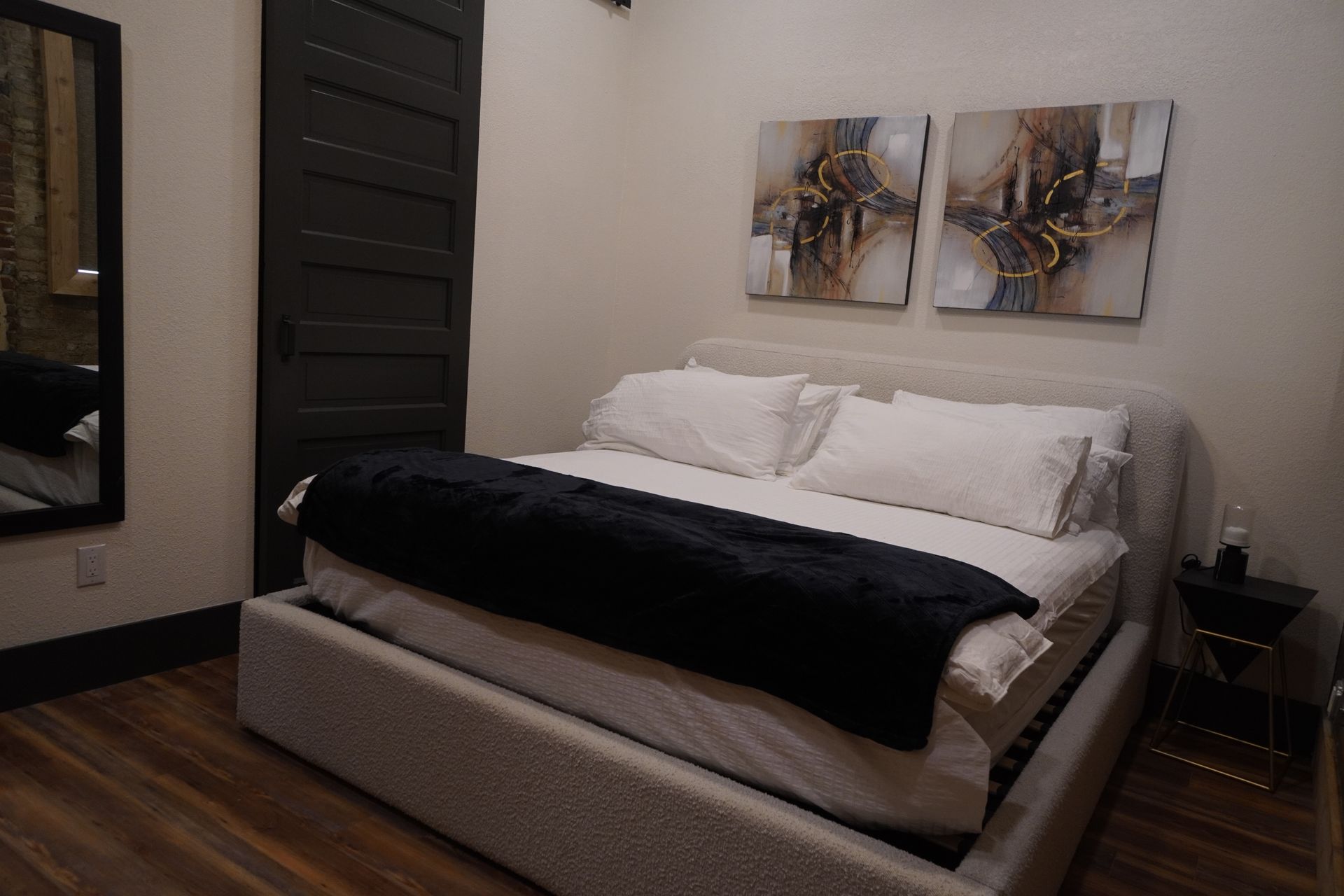 Bedroom with bed, art, black blanket, dark door and wood floors.