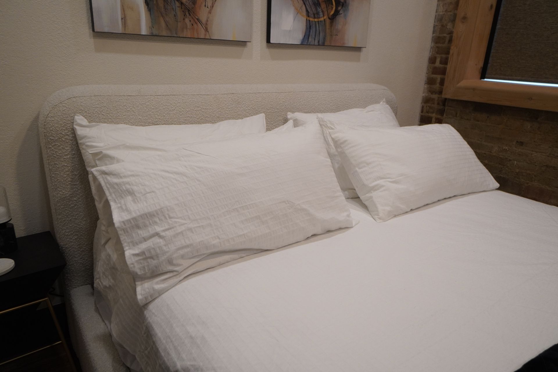 A bed with white bedding, pillows, and a textured headboard; brick wall in the background.