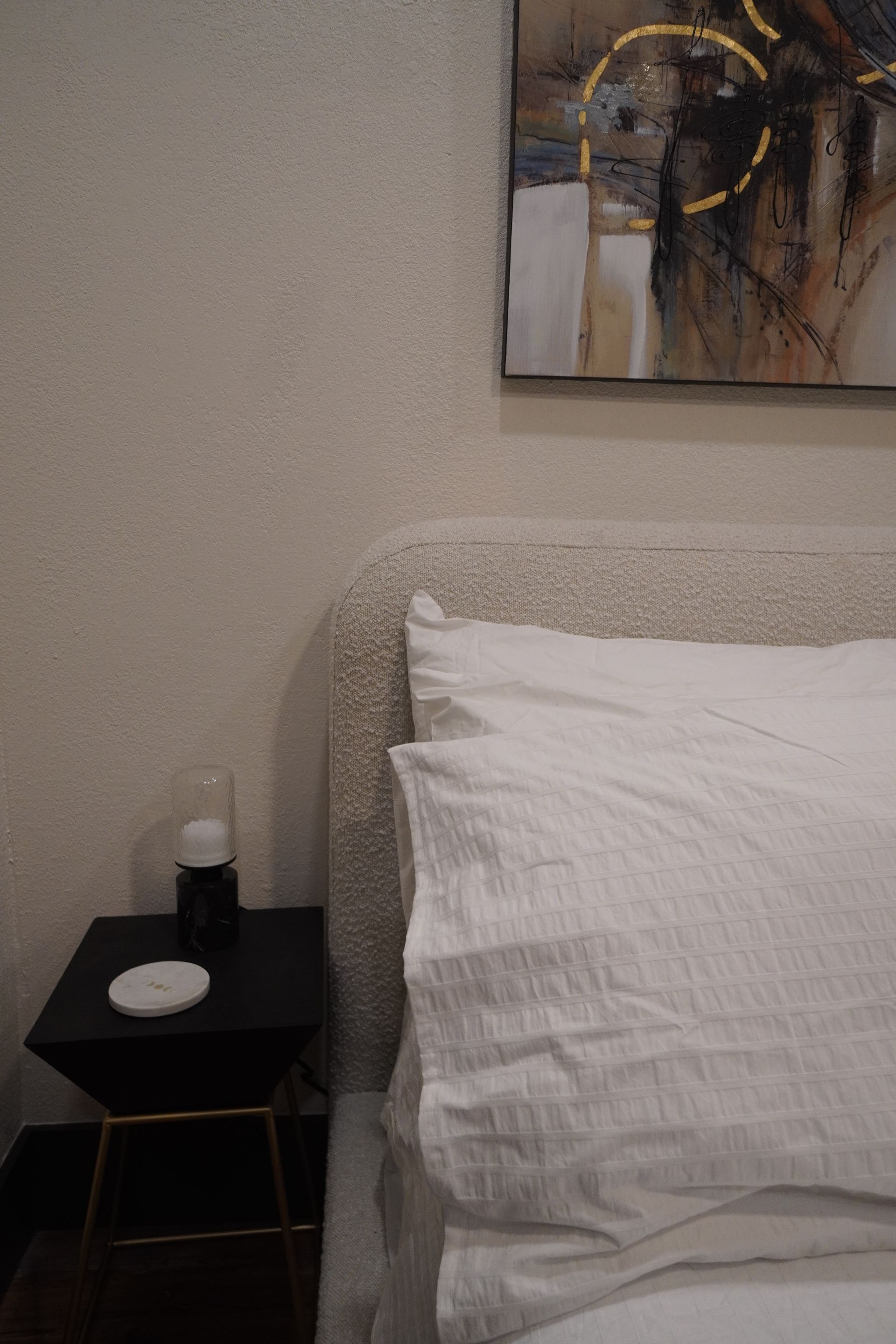 White bed with textured headboard, black nightstand, art print on the wall.
