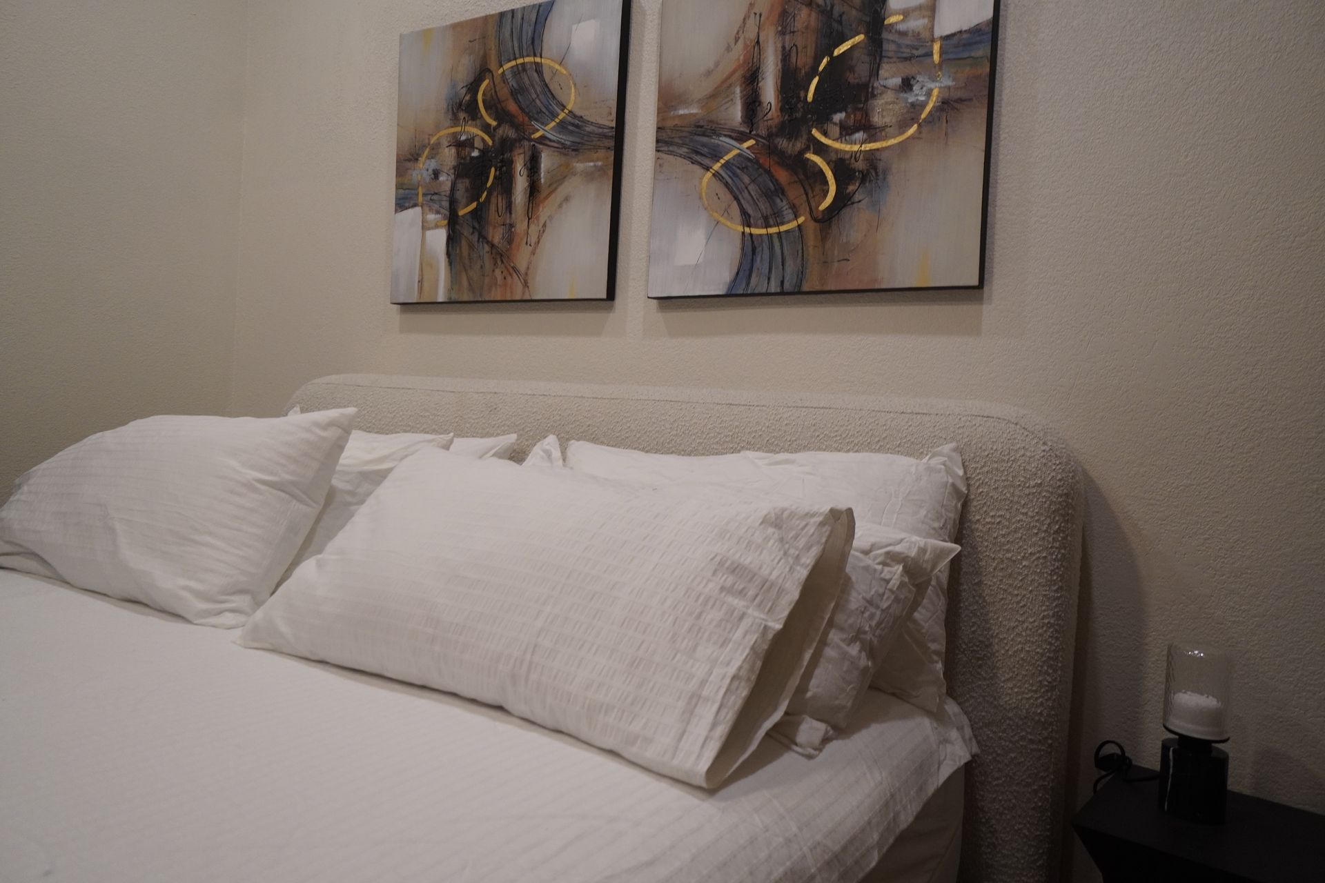 Bed with white linens, pillows, and textured headboard. Two abstract paintings hang above.