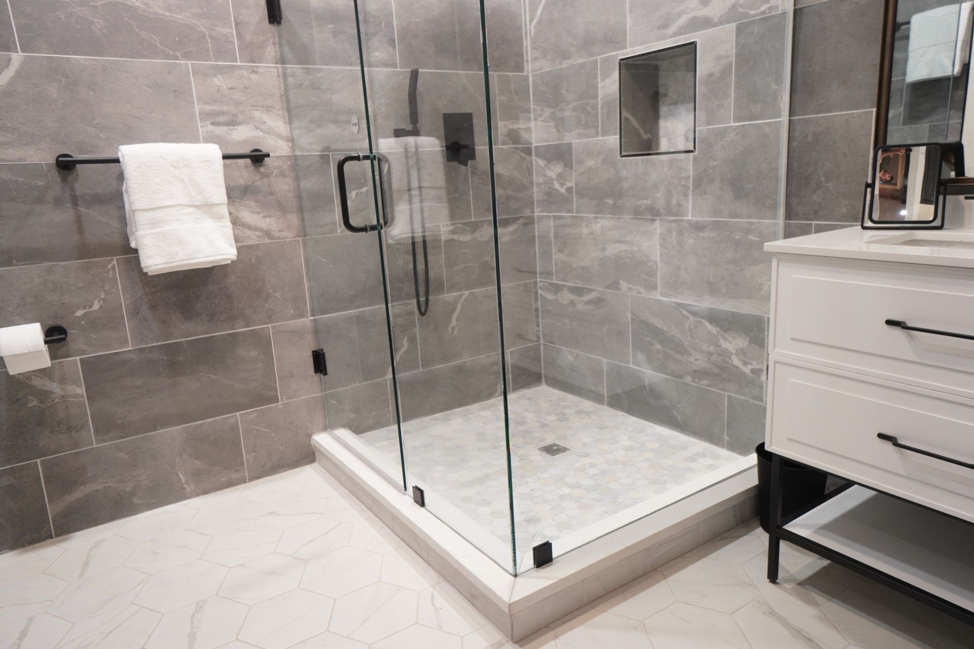 A modern bathroom with a glass shower, gray tiled walls, white vanity, and marble flooring.