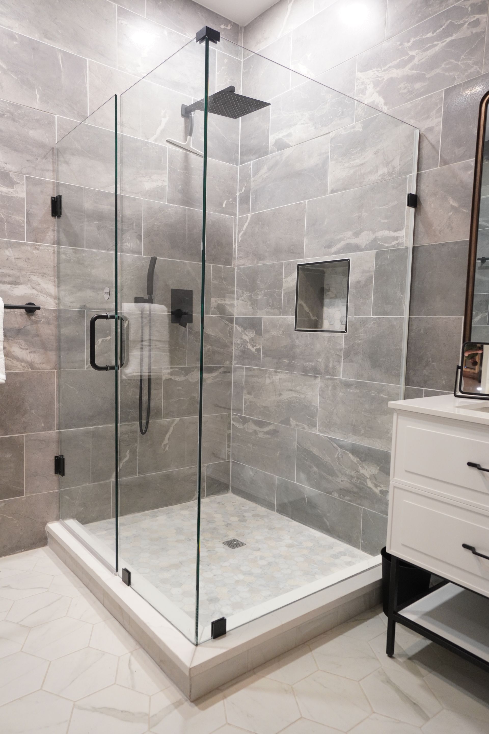 Modern bathroom with gray tiled walls, glass shower enclosure, and white vanity.