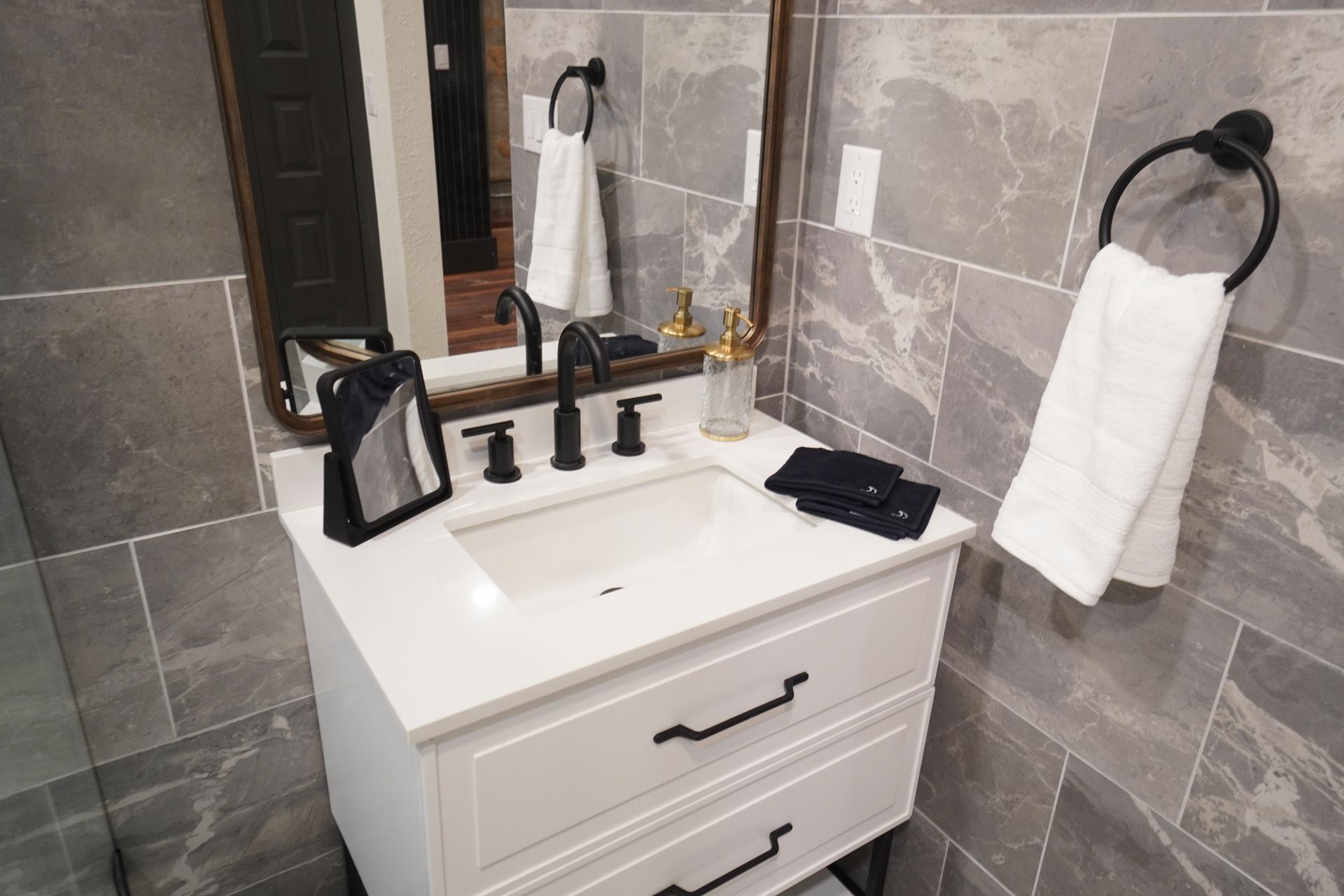 Bathroom vanity with white cabinet, gray tile, black fixtures, mirror, and towel.