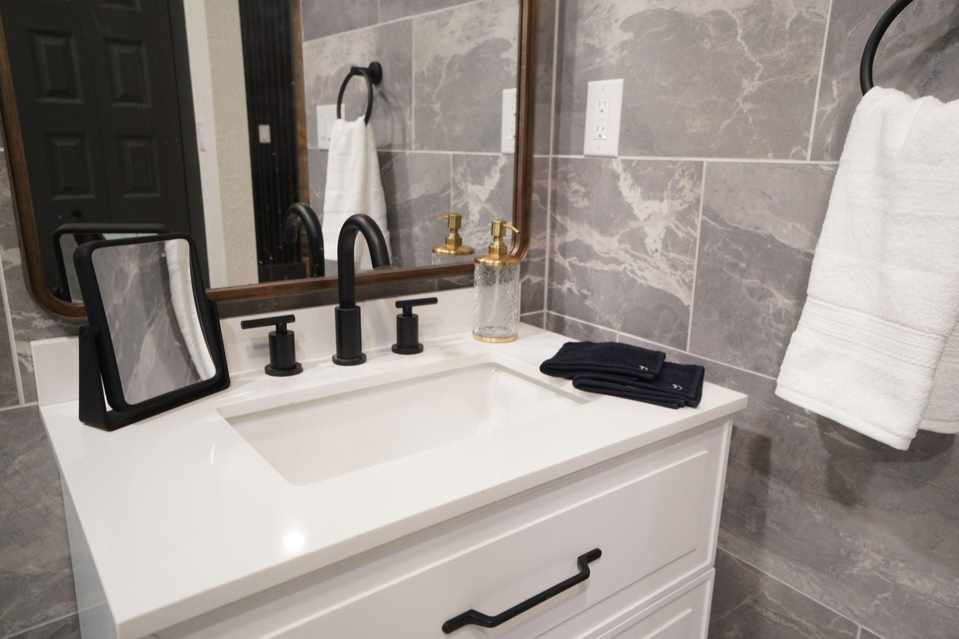 Bathroom vanity with a white countertop, black fixtures, and gray tiled walls.