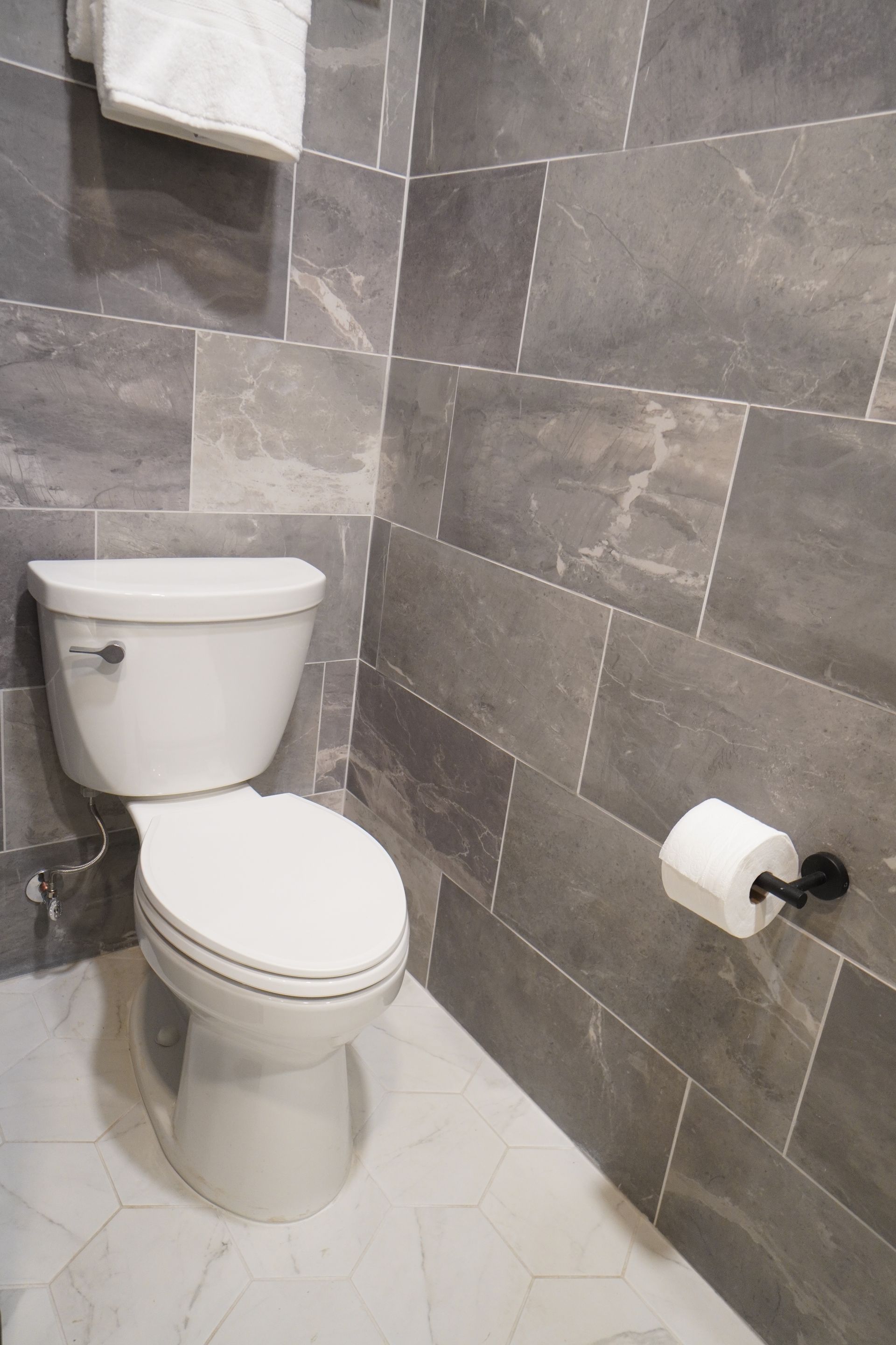 White toilet in a bathroom with gray tiled walls and a roll of toilet paper.