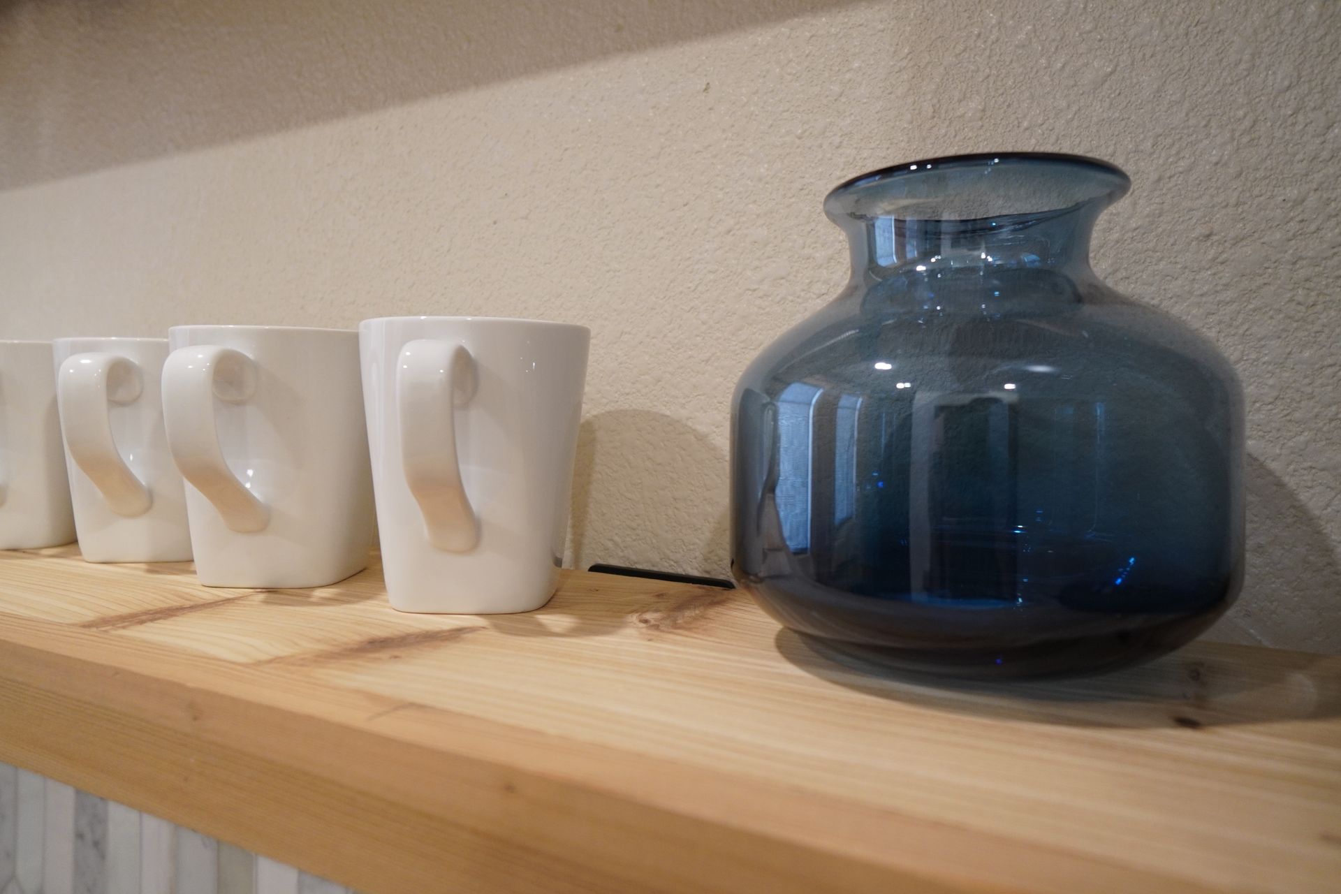White mugs and blue vase on a wooden shelf against a beige wall.