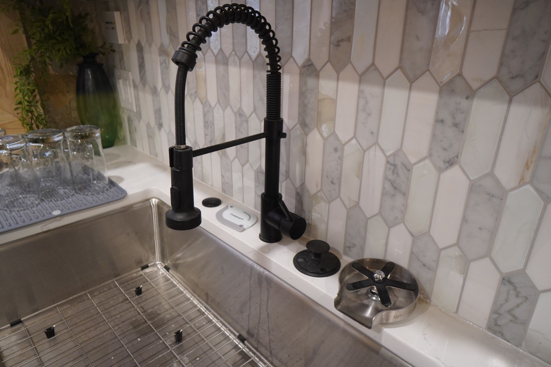 Stainless steel kitchen sink with black faucet, white countertop, and patterned tile backsplash.