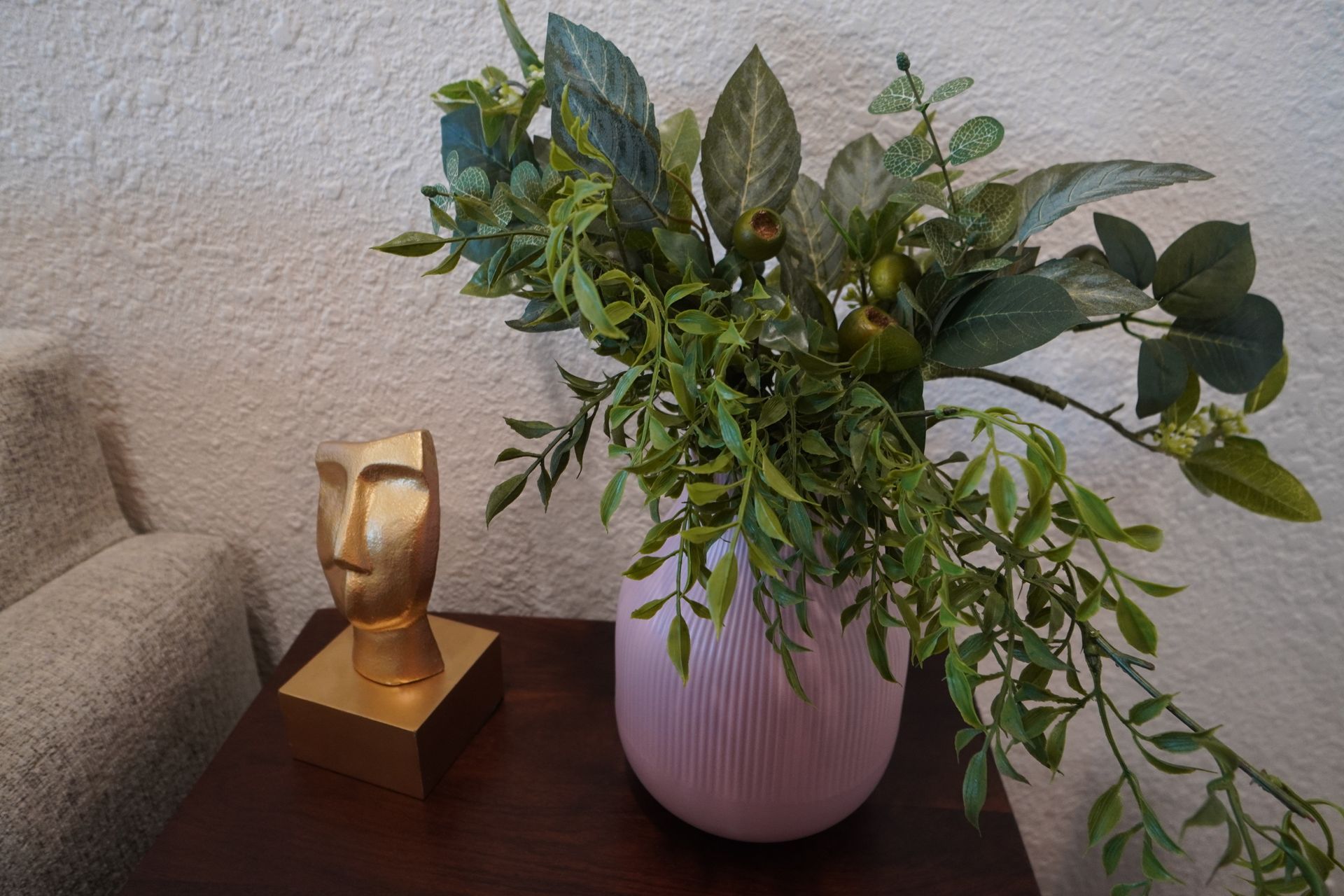 Pink vase with green leaves and golden face sculpture on a dark brown table.