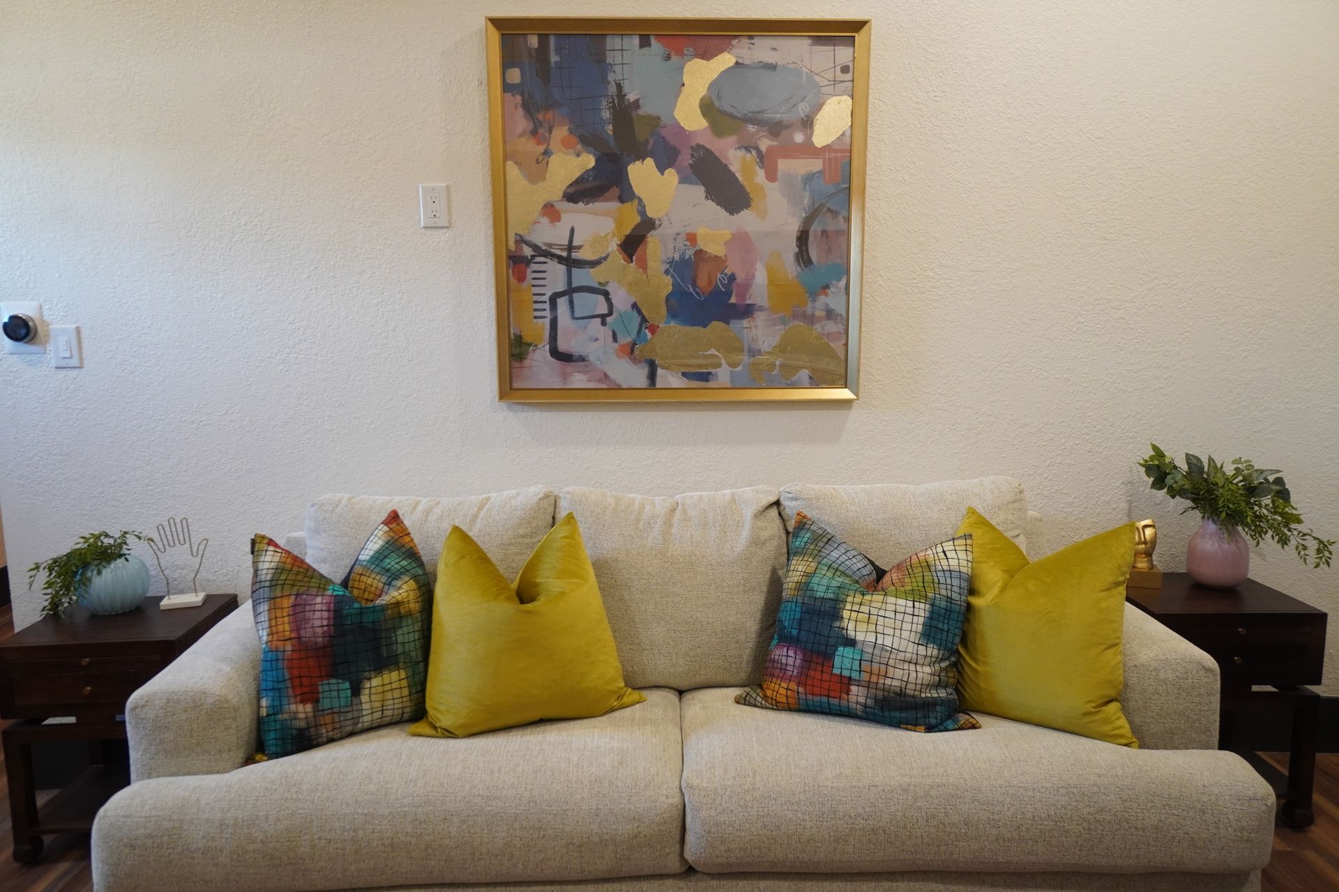 Cream couch with colorful pillows beneath abstract art; two dark side tables with plants on each side.