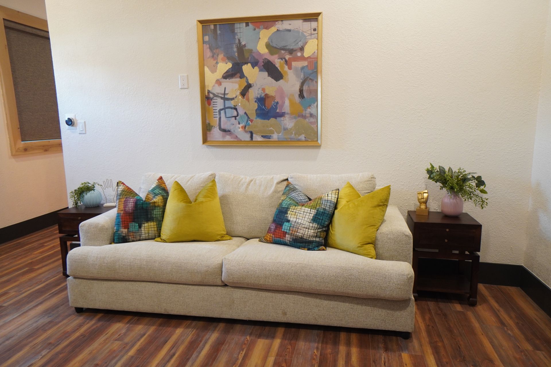 A cream sofa with colorful pillows beneath abstract art on a white wall; flanked by dark wood end tables.