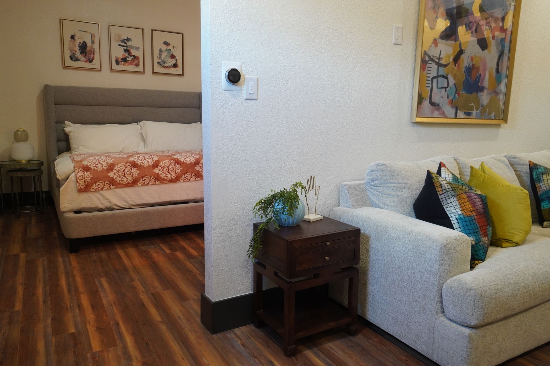 Bedroom view with bed and side table next to sofa and art, warm lighting, and hardwood floors.
