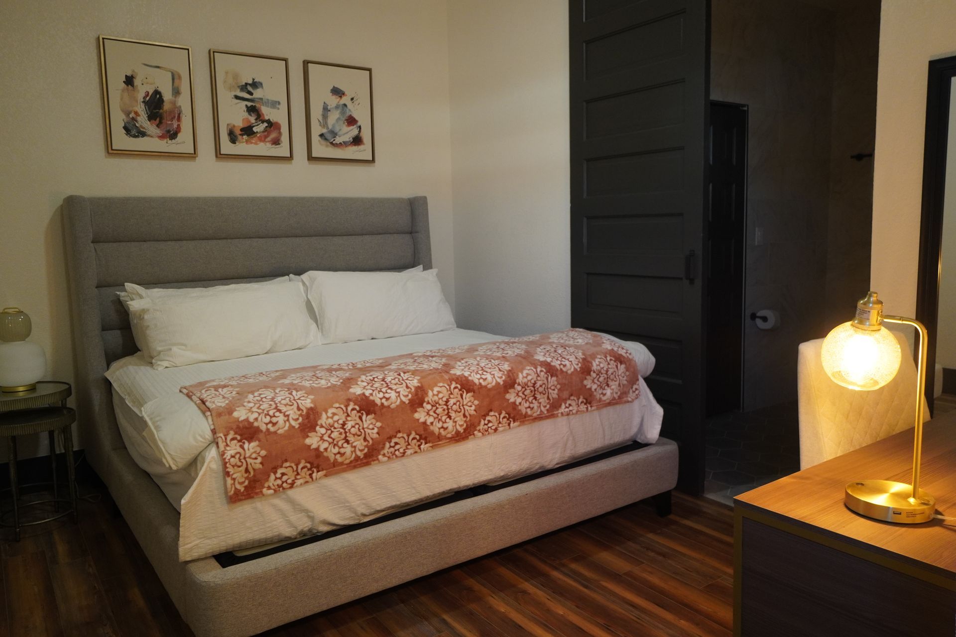 Bedroom with gray bed, peach blanket, art prints, dark door, wooden desk, and lit lamp.