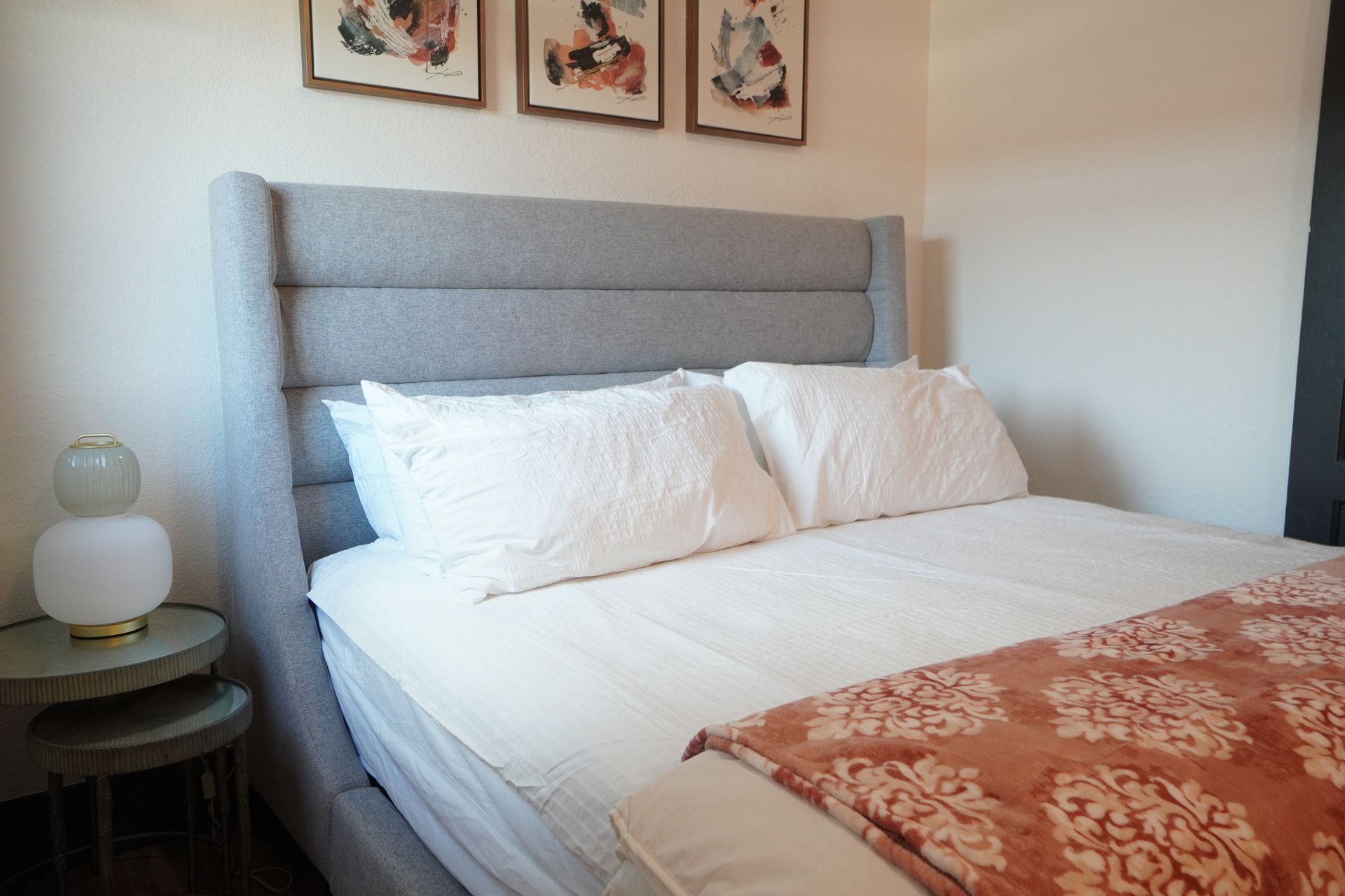 A bed with a gray headboard, white bedding, and a patterned orange throw. Artwork hangs above.