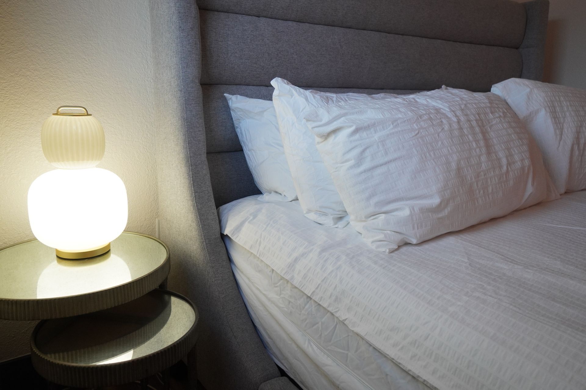 Bedside table with lamp next to a bed with white pillows and bedding.