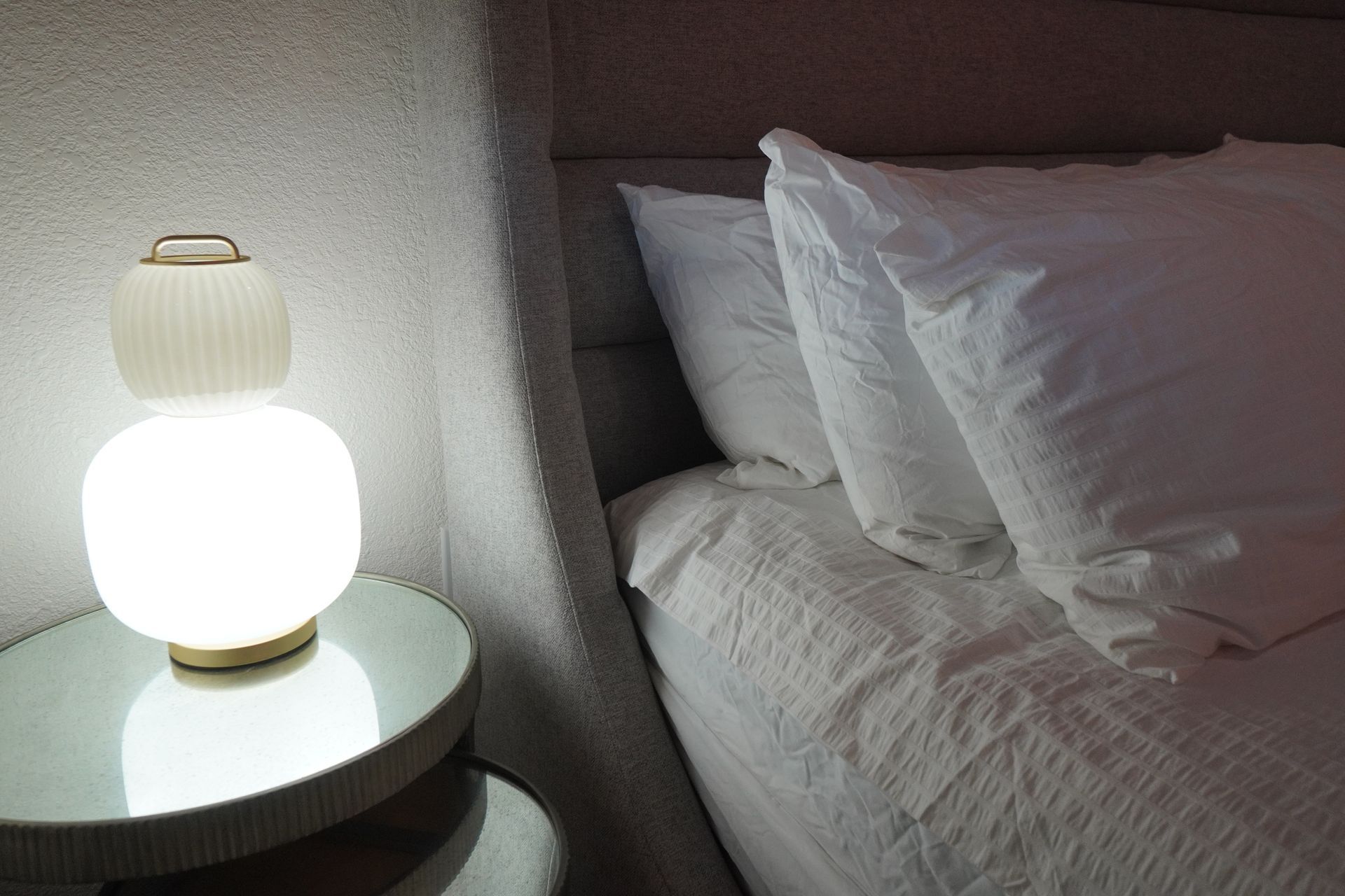 A glowing bedside lamp sits on a mirrored table beside a bed with white pillows and bedding.