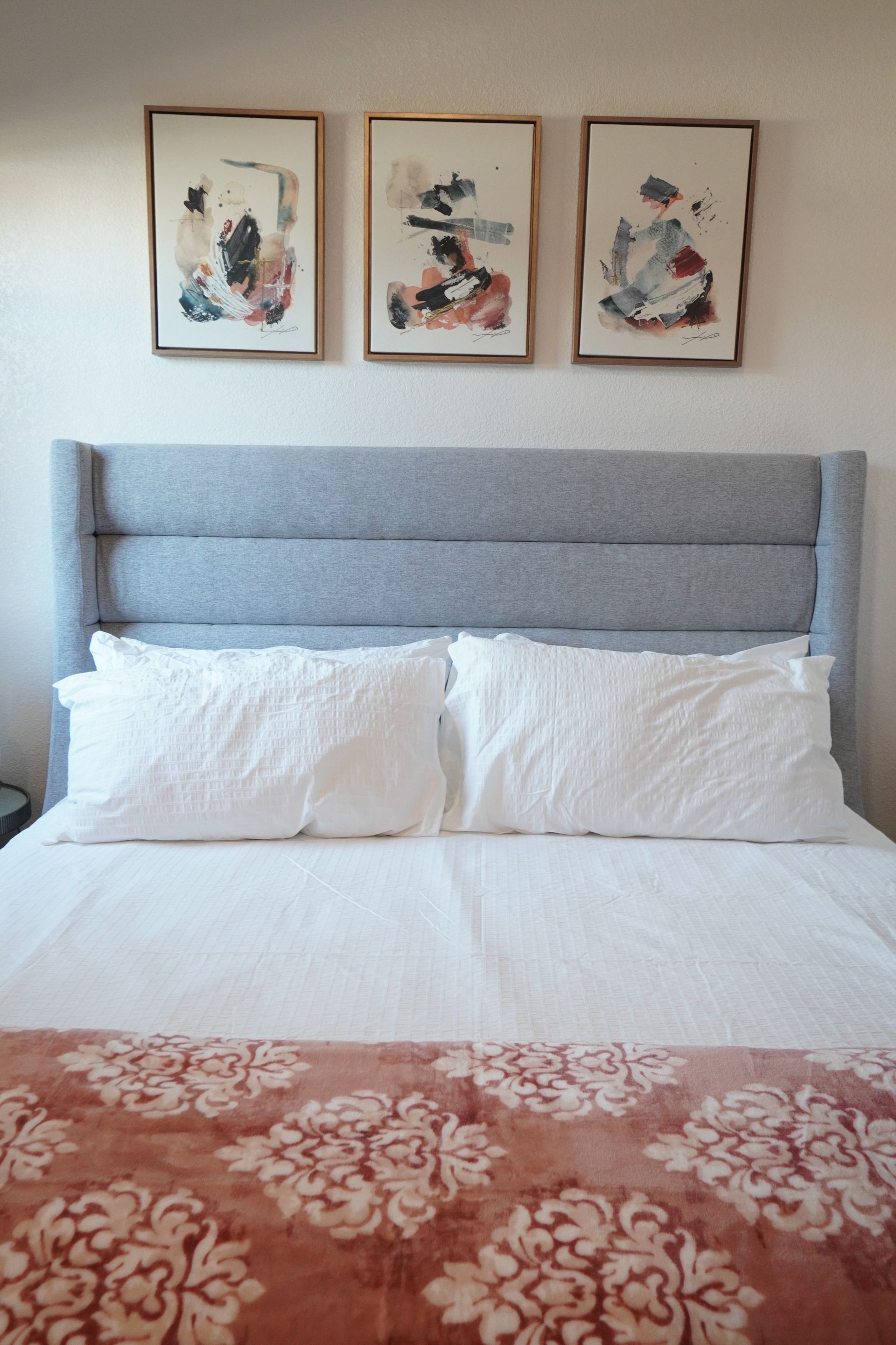 Bed with gray headboard, white bedding, and three framed abstract art pieces above. 