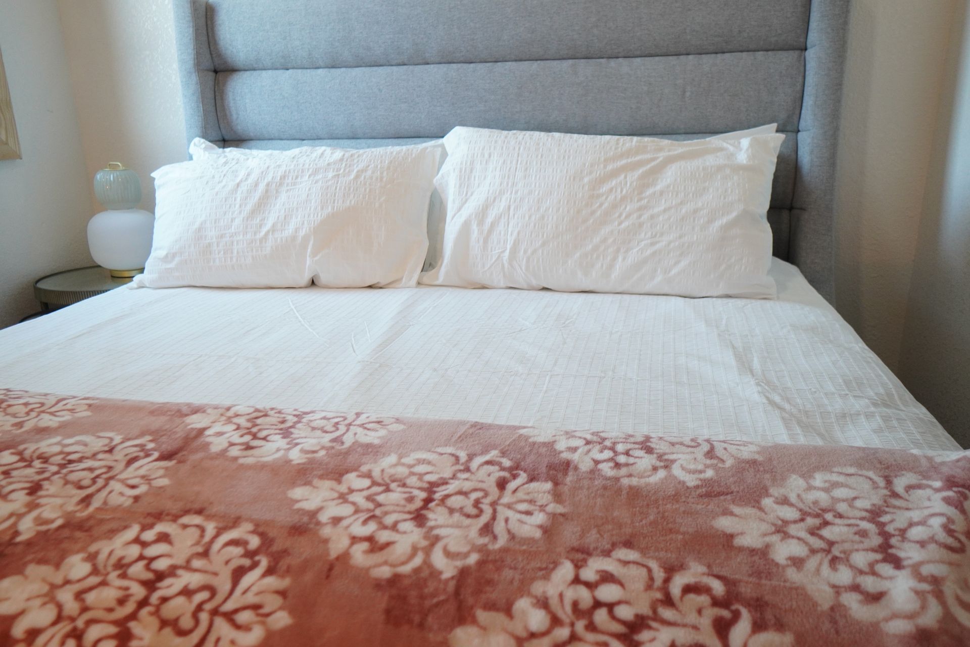 Bed with white bedding, two pillows, pink floral blanket, and gray headboard.