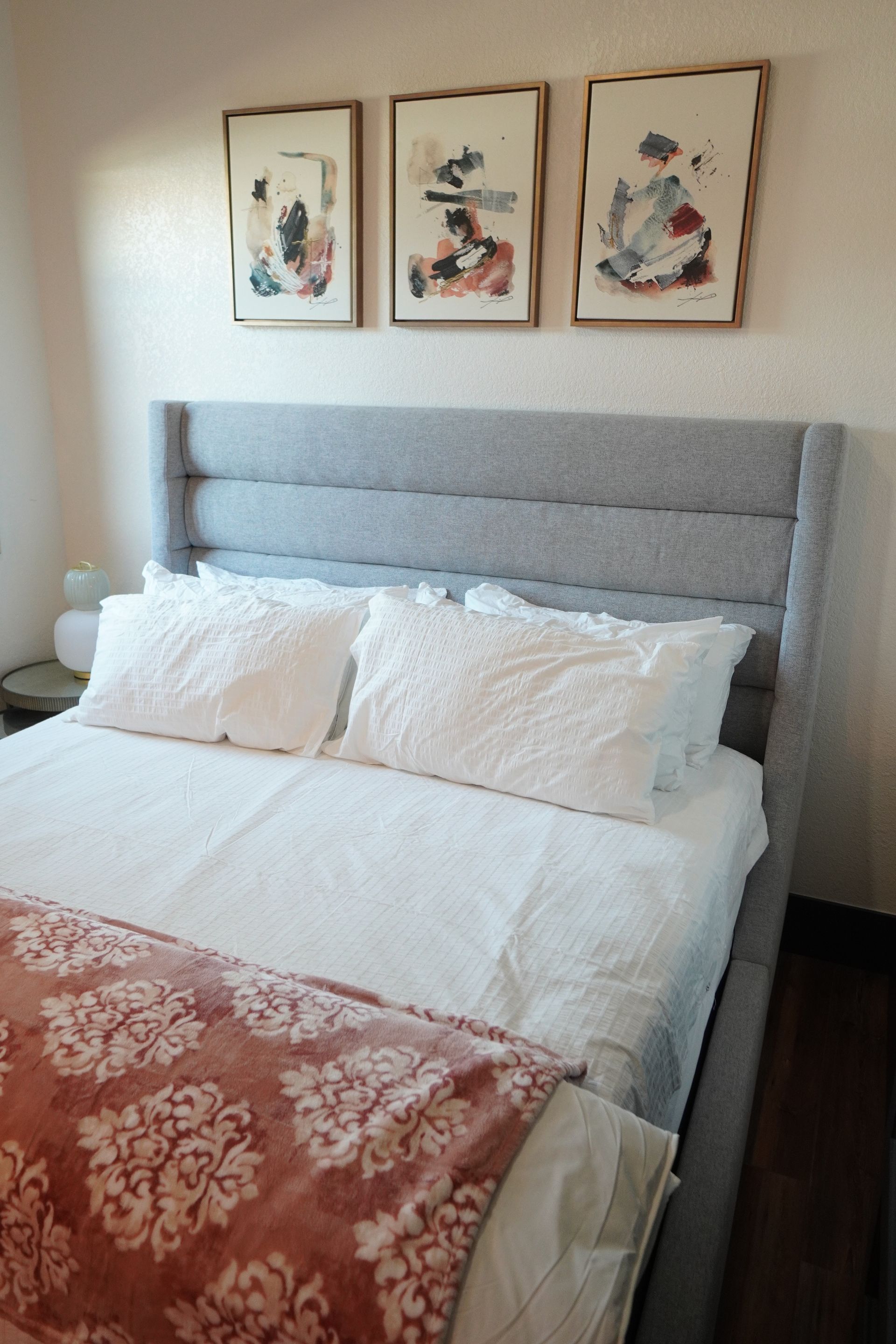 Bedroom with a gray upholstered bed frame and headboard, white bedding, and three abstract artworks above.