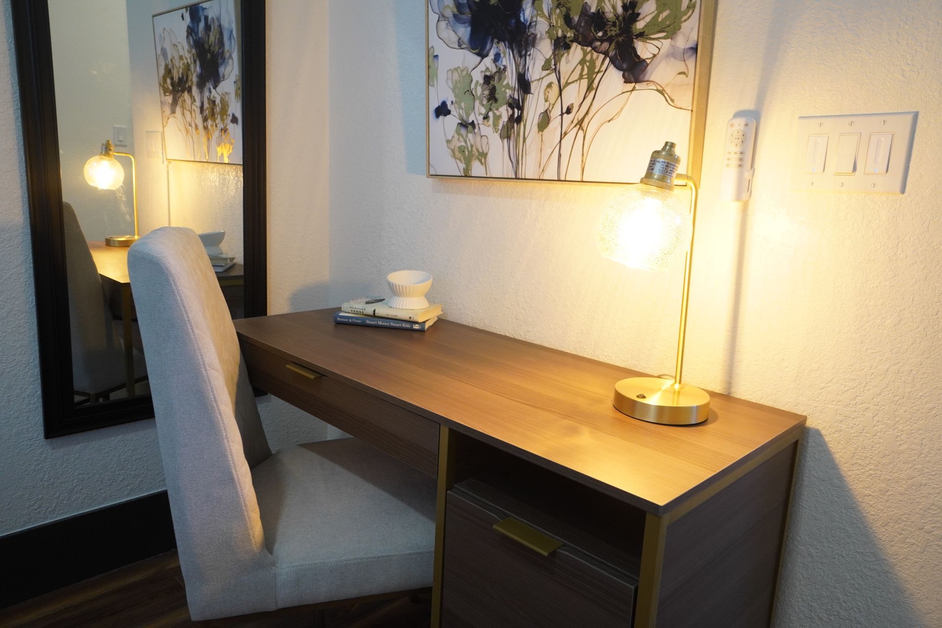 Desk with lamp, mirror, and chair. Artwork hangs on the wall. Well lit corner.