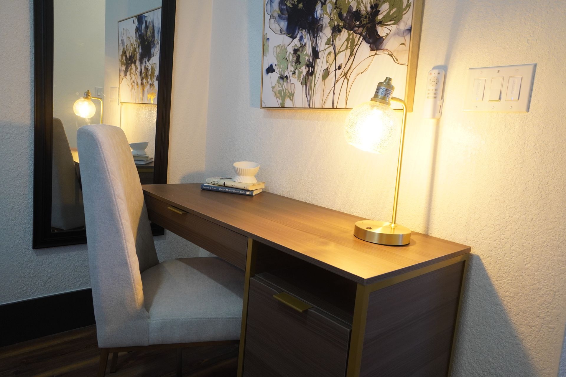 Desk with chair, lamp, and mirror in a room. Wood desk with a drawer and cabinet, light-colored chair.