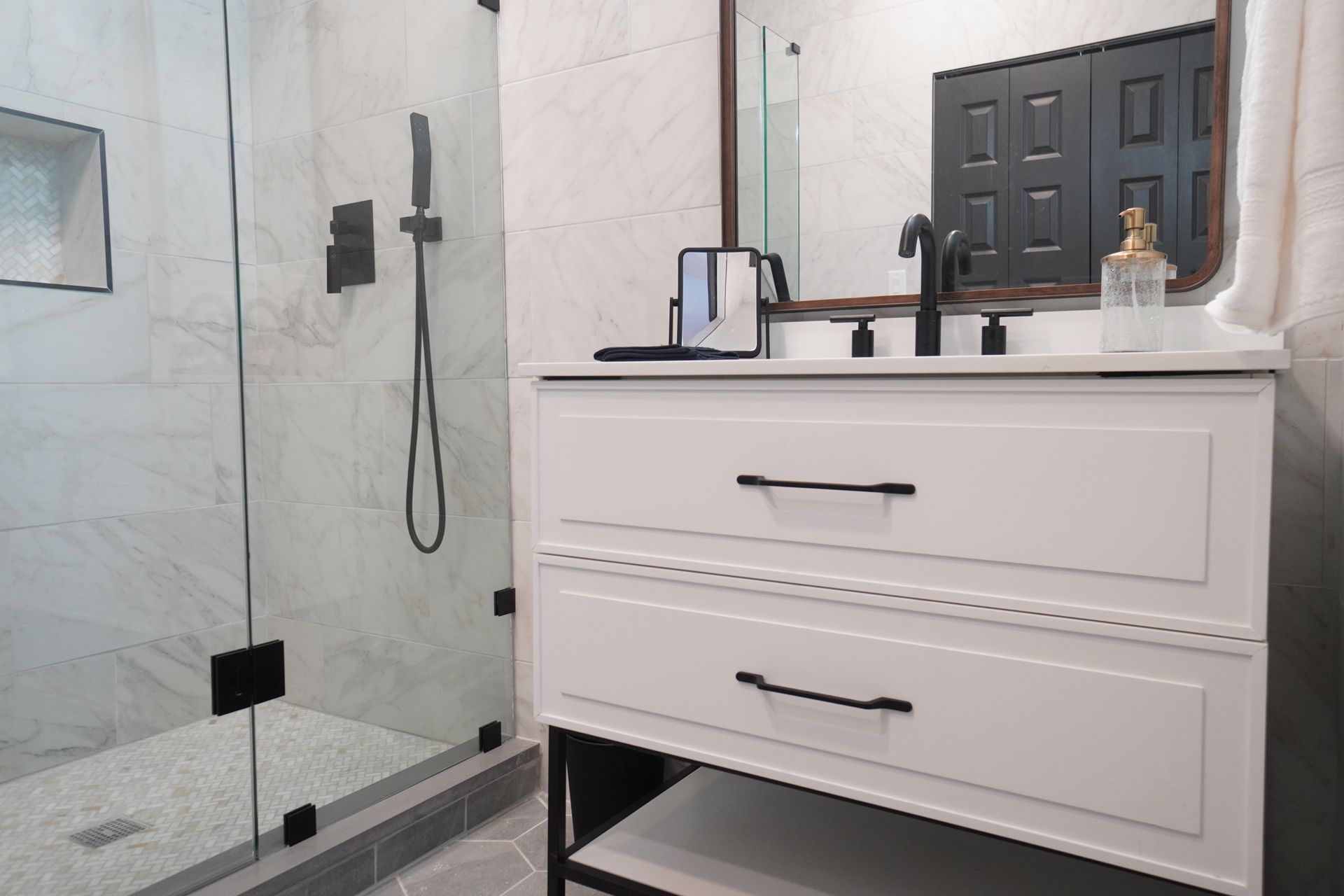 White bathroom vanity with black hardware, next to a glass shower with black fixtures.