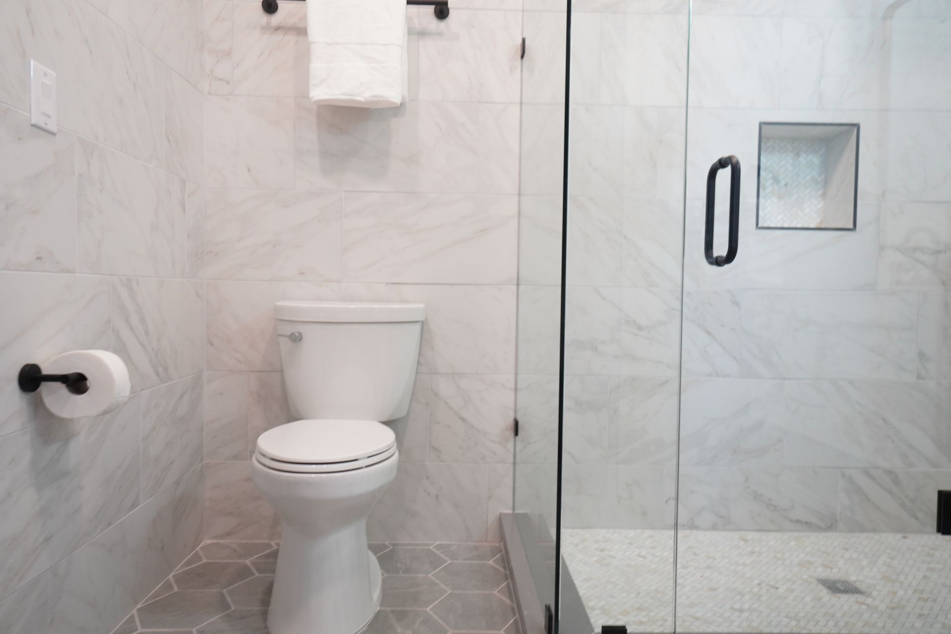 Modern bathroom with white marble tile, toilet, glass shower, and black fixtures.
