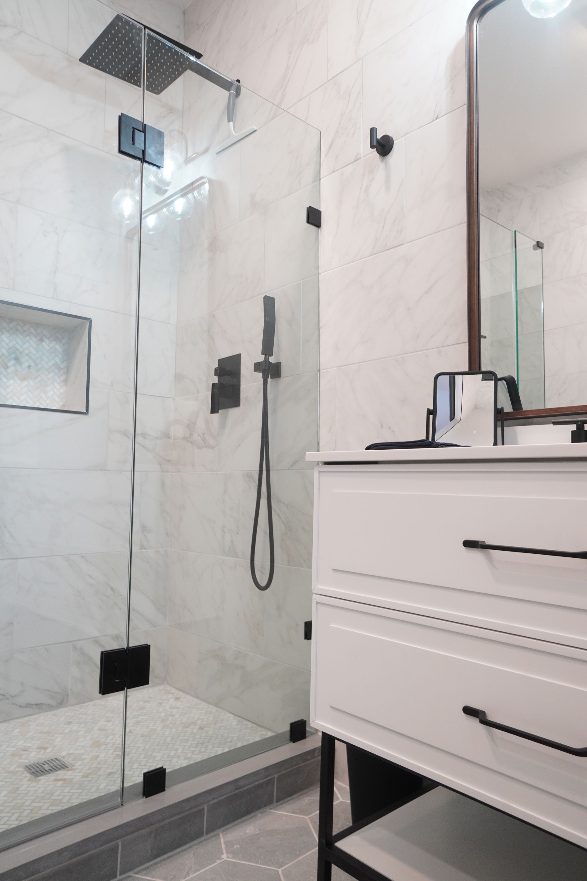 Modern bathroom with glass shower, white vanity, dark hardware, and light-colored tile.