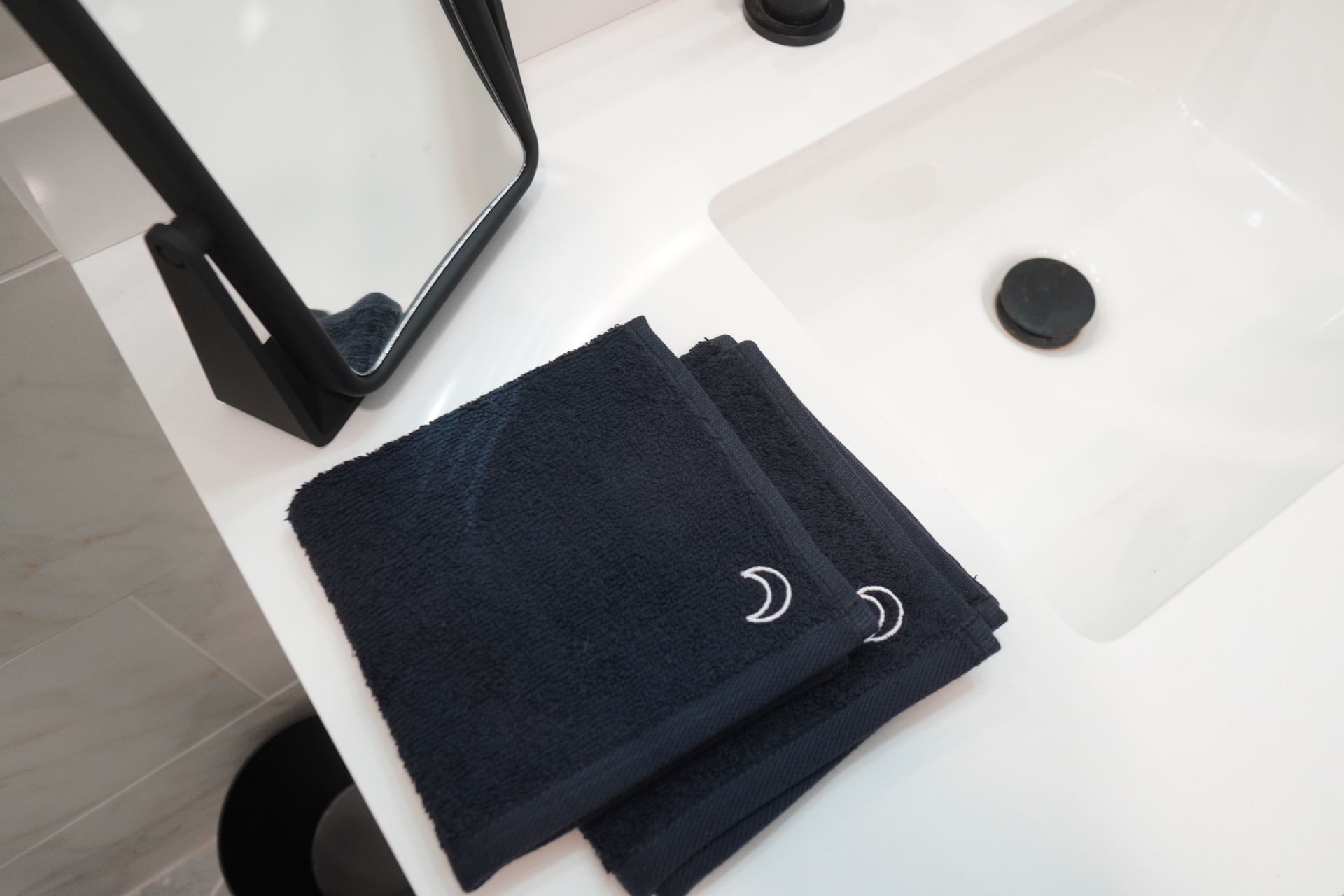 Dark blue washcloths with silver crescent moon detail on a white countertop by a sink and mirror.