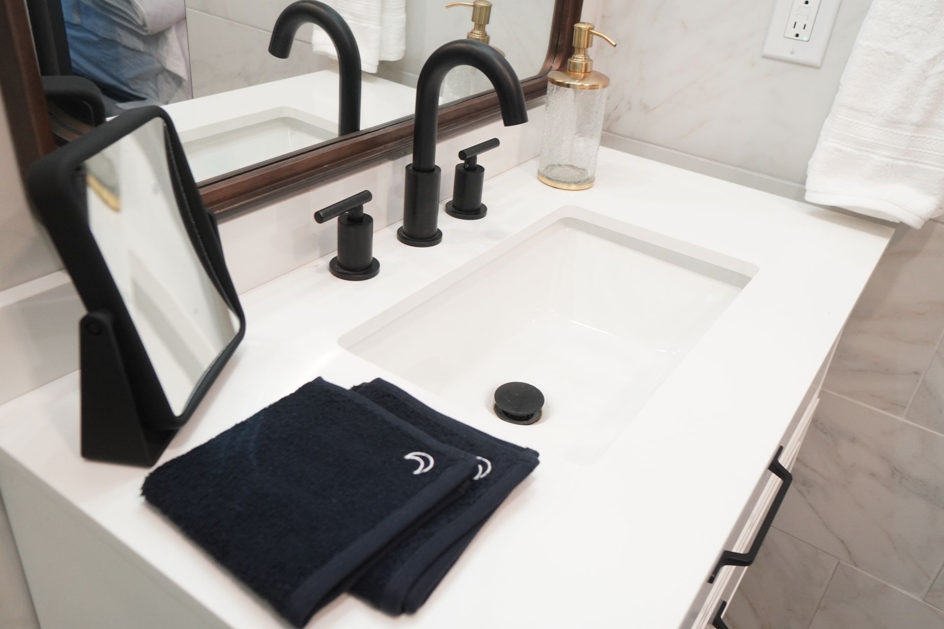 White bathroom countertop with sink, black faucet, mirror, towels, and soap dispenser.