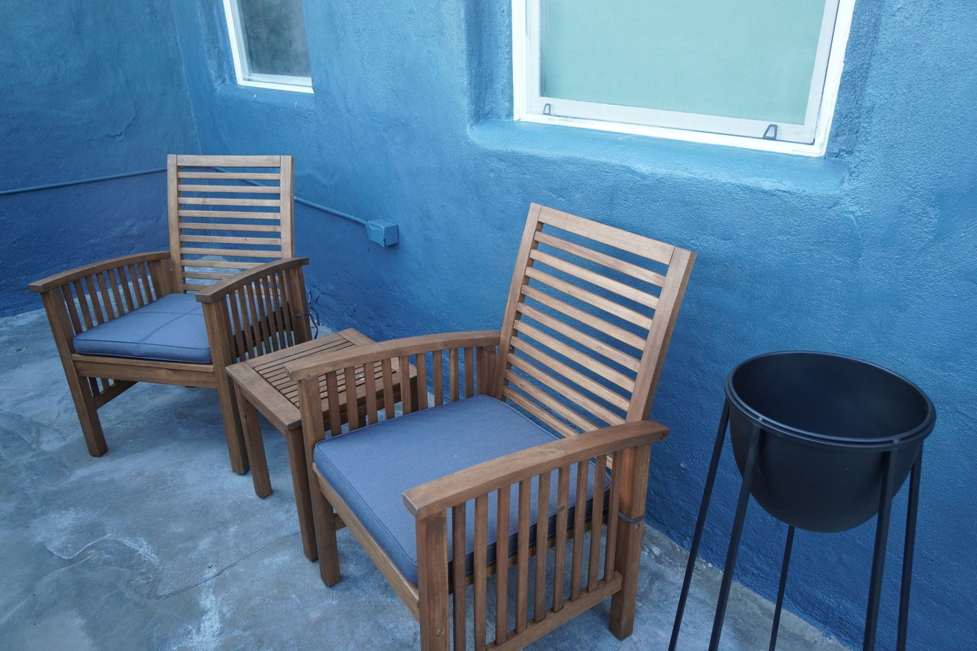 Two wooden chairs with blue cushions and small table on a blue patio near a window and plant stand.