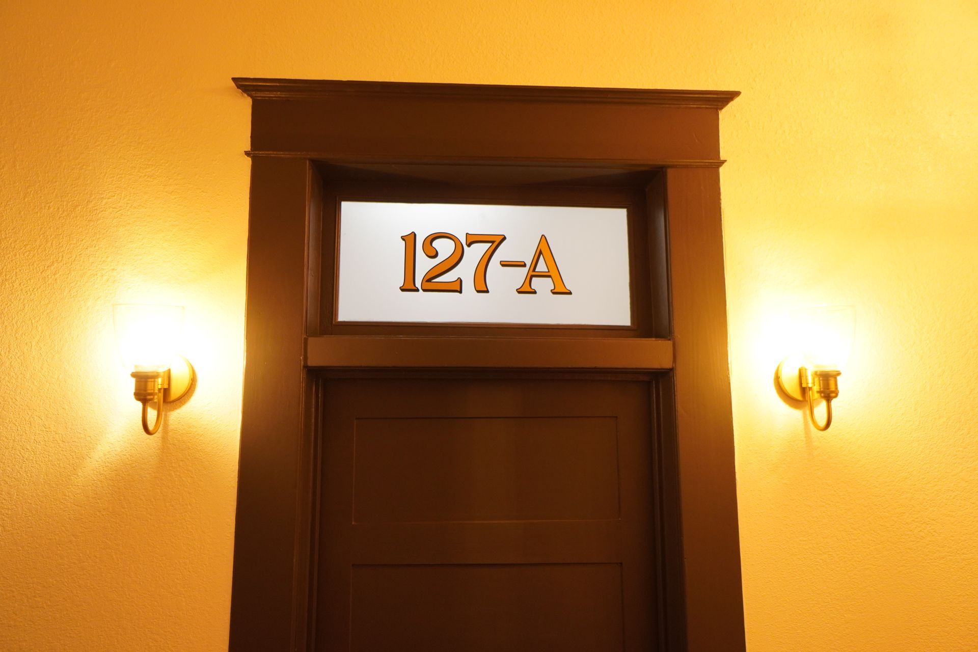 Door with number 127-A above, flanked by sconce lights on a golden wall.