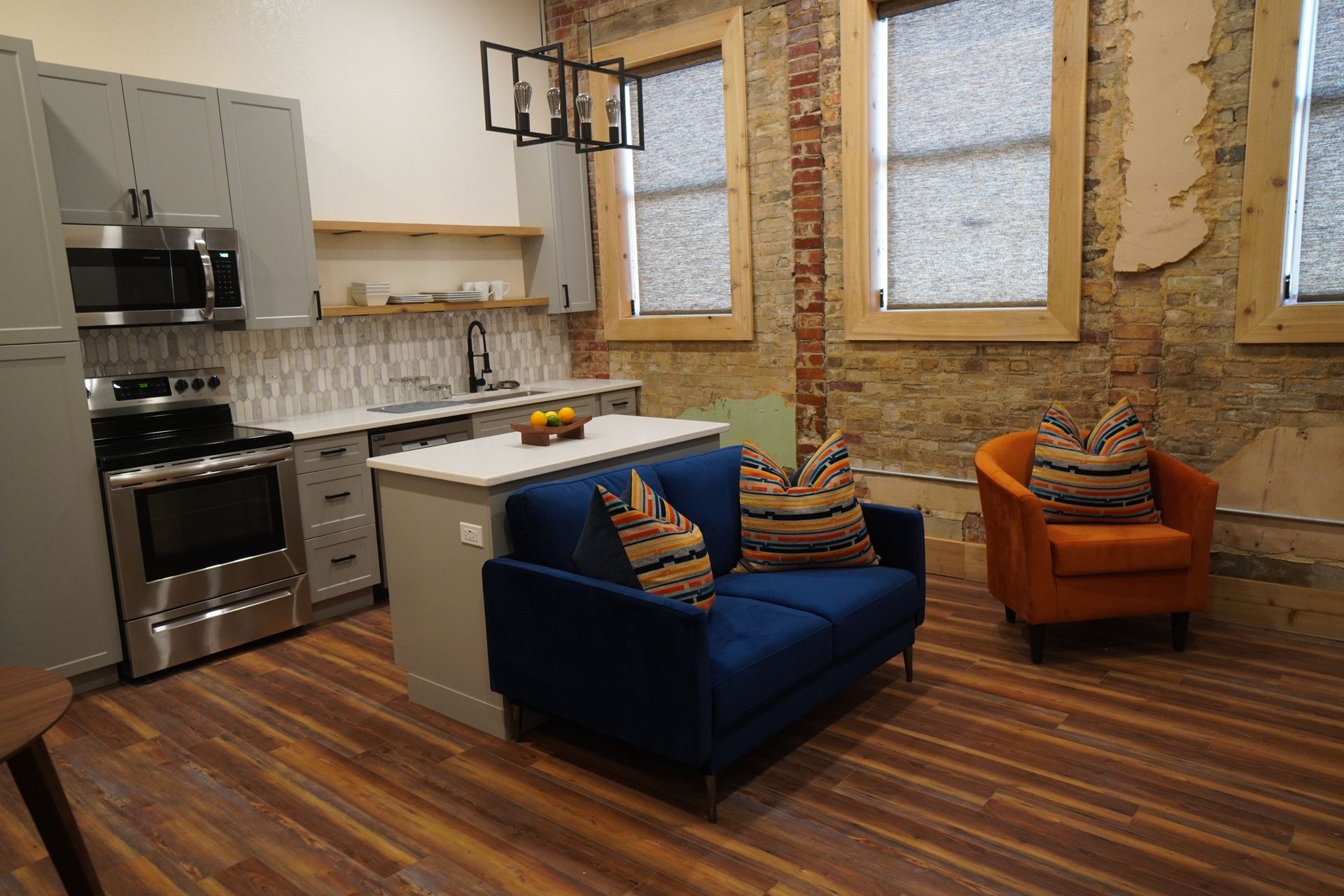 Modern apartment kitchen with blue loveseat and orange armchair against exposed brick wall.