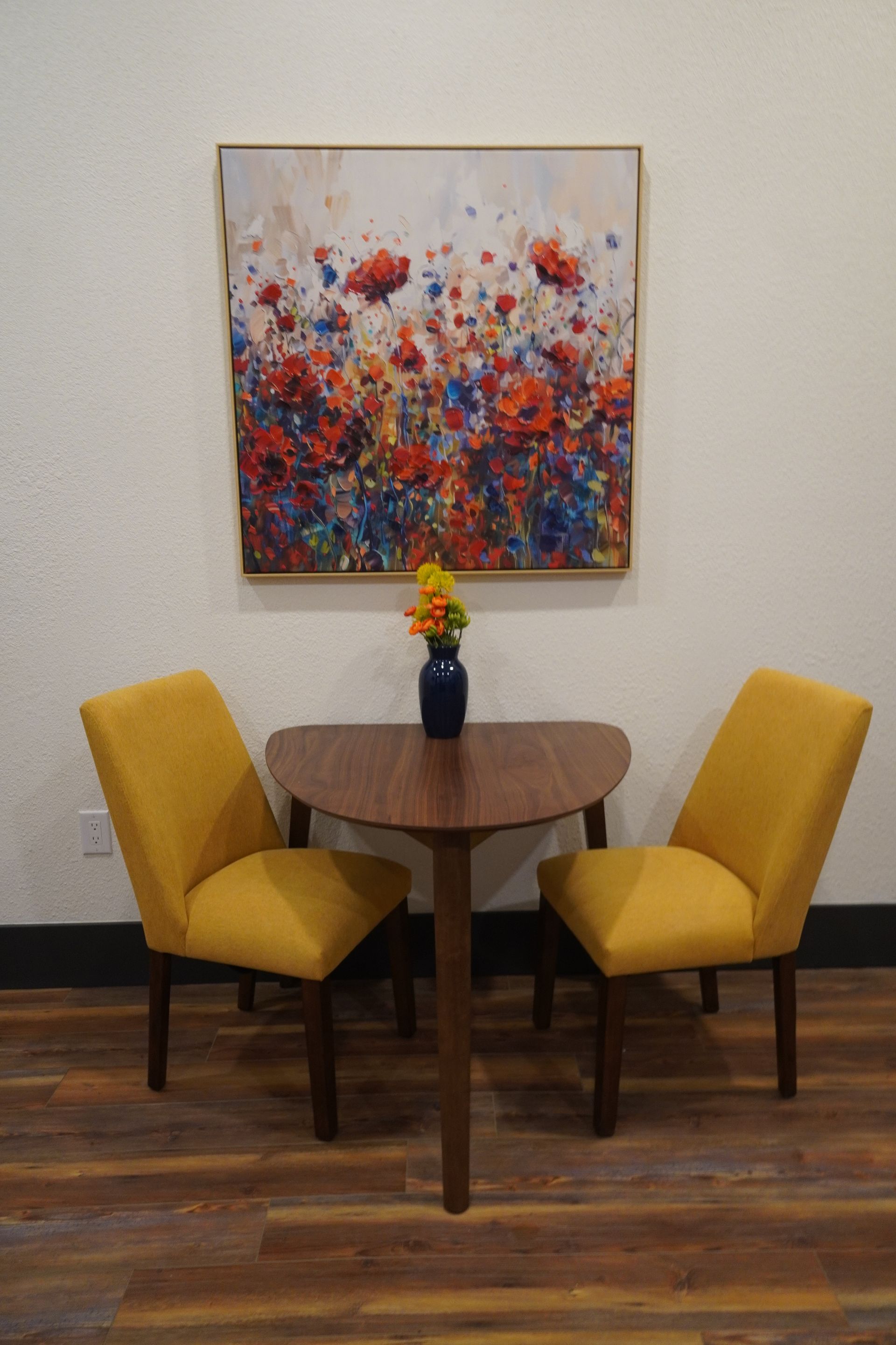 Dining area with small wooden table, two yellow chairs, floral painting, and vase of flowers.