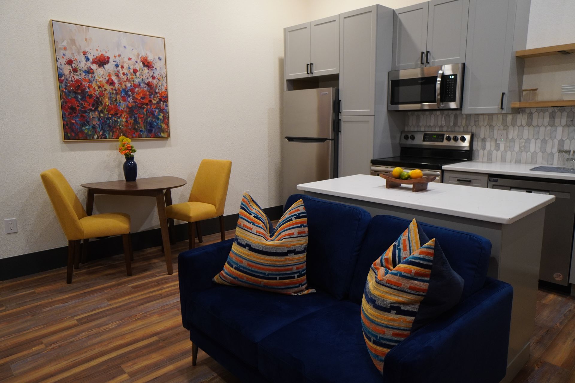 Cozy open-plan living space with blue sofa, dining table, and small kitchen with gray cabinets.